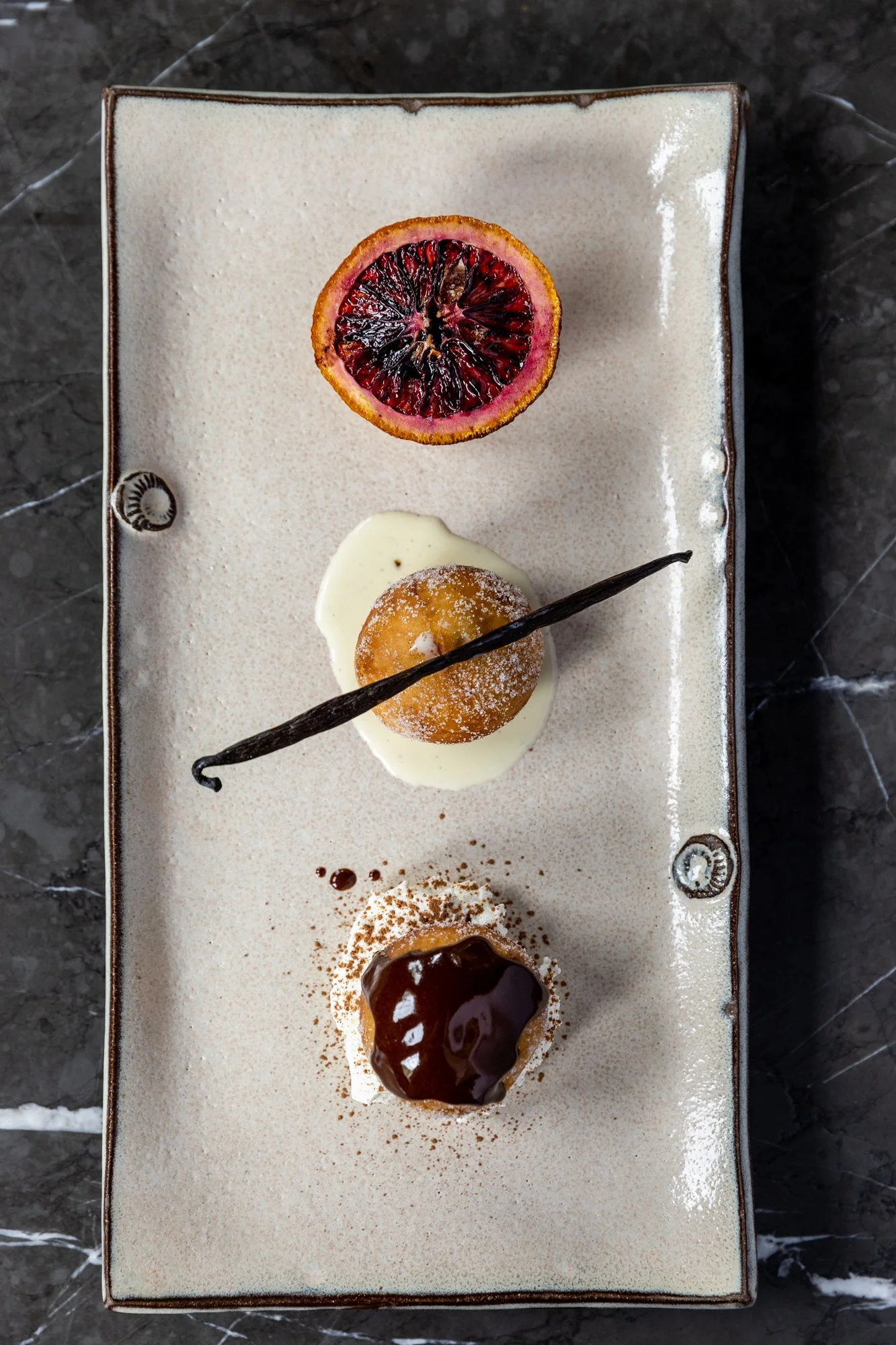 A rectangular ceramic plate with three dessert items: a halved blood orange at the top, a small round pastry with white glaze and a vanilla bean on top in the middle, and a chocolate-covered profiterole with cocoa powder at the bottom on a dark marbl