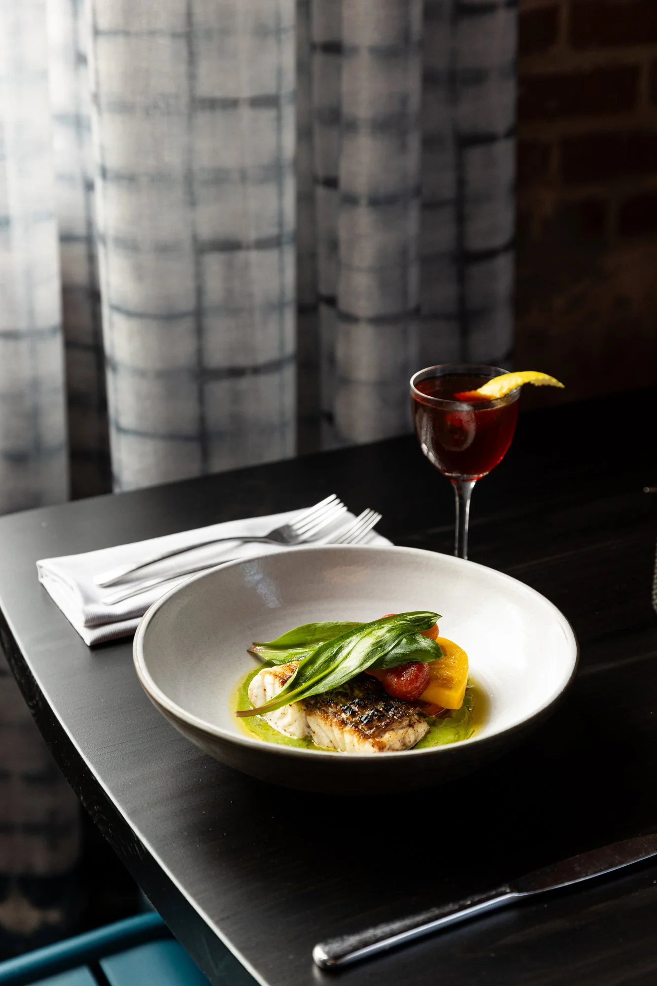 A gourmet fish dish garnished with green herbs and vegetables, served in a white bowl with a lemon wedge and accompanied by a glass of red wine with a lemon garnish, on a dark wooden table with a white napkin and silverware.