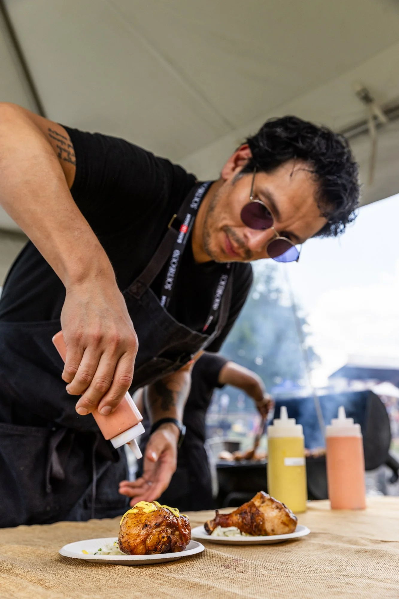 A man with curly black hair, wearing sunglasses and a black shirt, is dressed in a black apron and is squeezing sauce onto grilled chicken drumsticks at an outdoor event, with condiments in the background. Southbound Food Festival in Birmingham, Alab