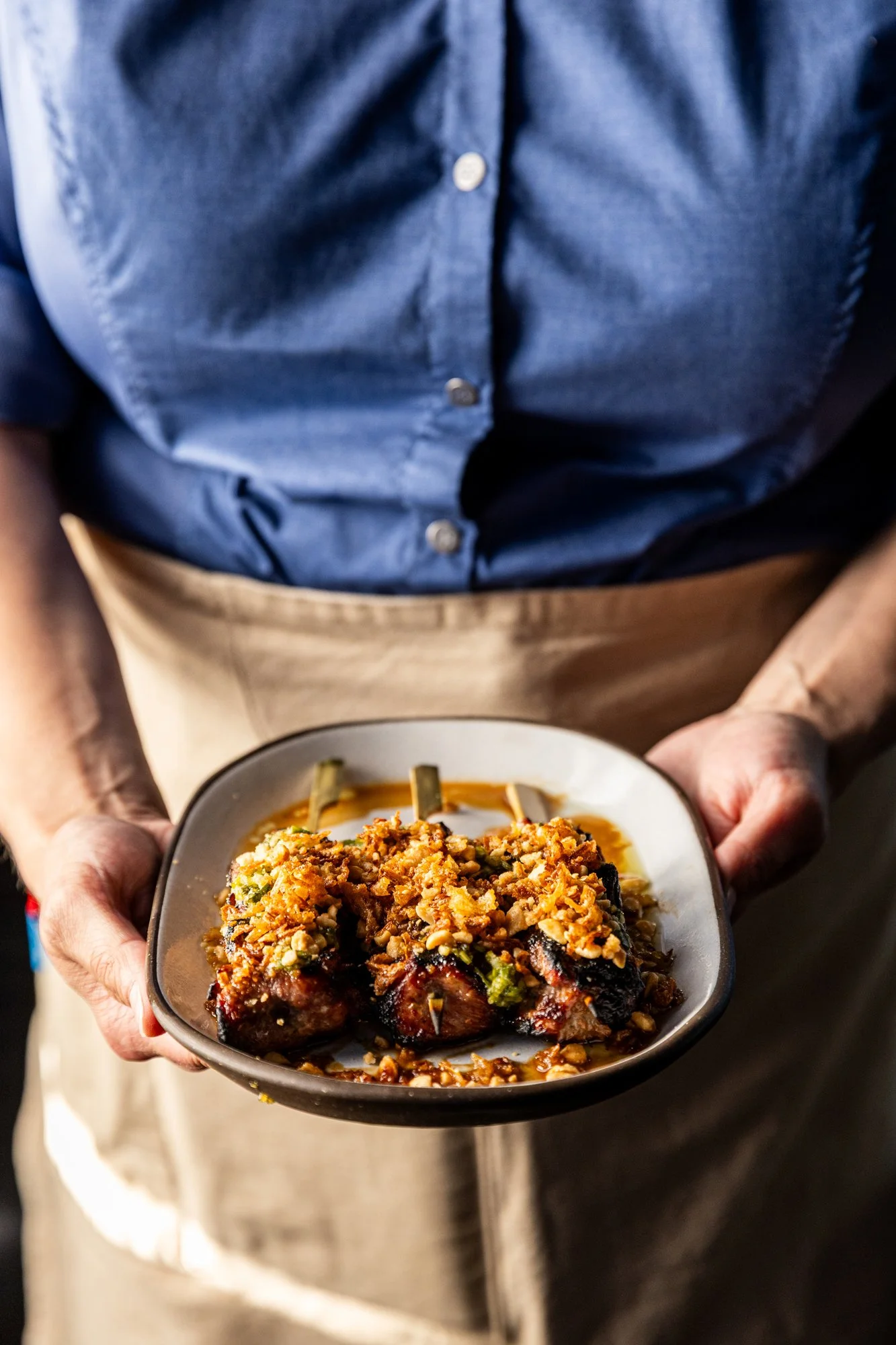 Person wearing a blue shirt and beige apron holding a bowl of glazed pork ribs topped with chopped nuts and herbs.