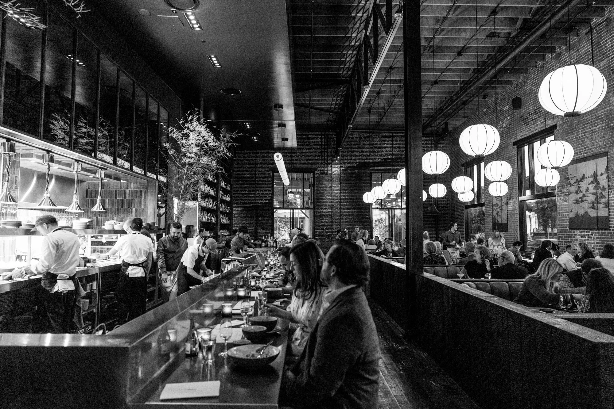 Black and white photo of a busy restaurant with patrons dining at tables and chefs preparing food behind a counter, illuminated by hanging paper lanterns.
