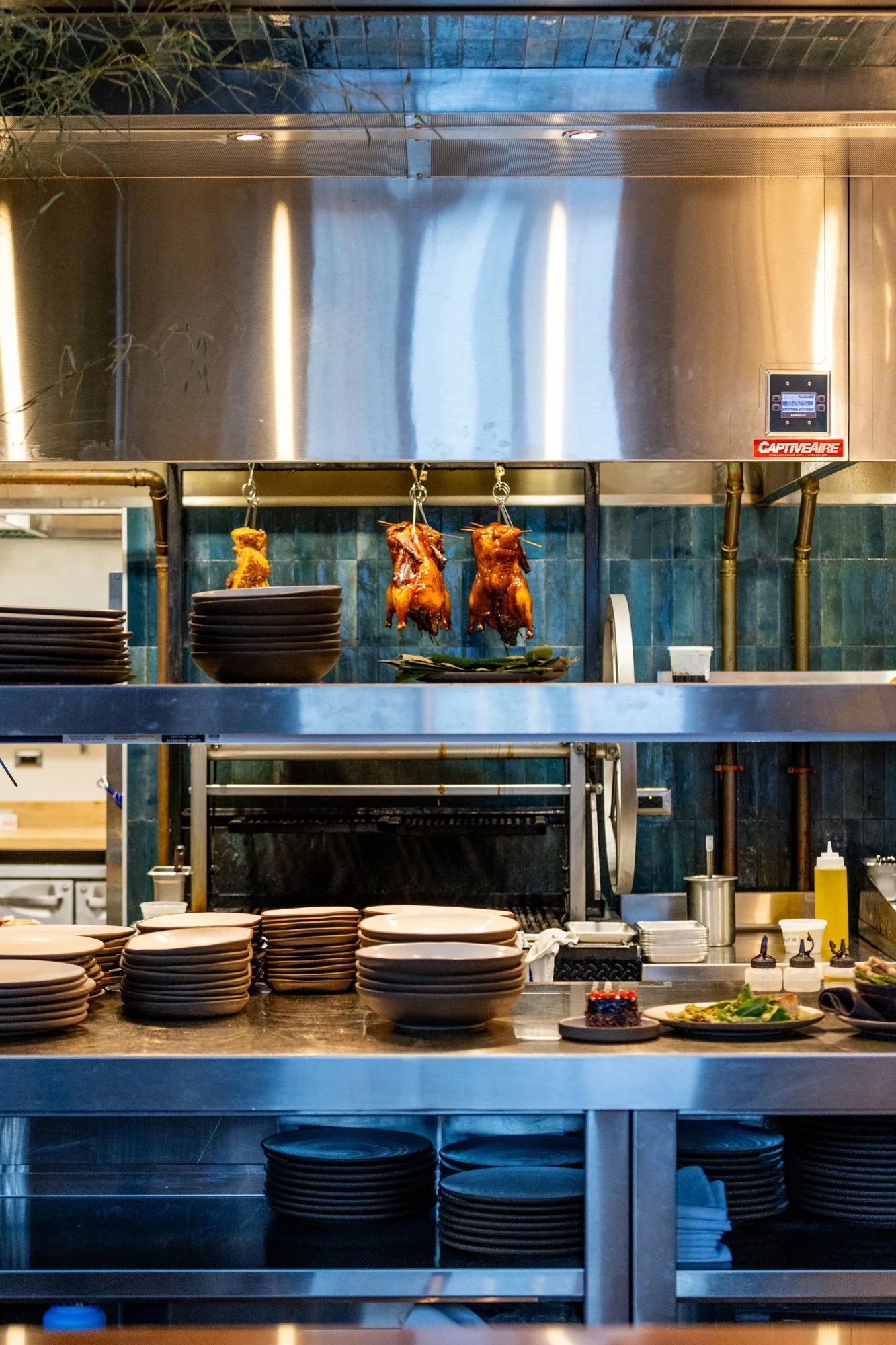 Kitchen with hanging roasted ducks, stacked black plates, and a countertop with plates, sauces, and a dessert.