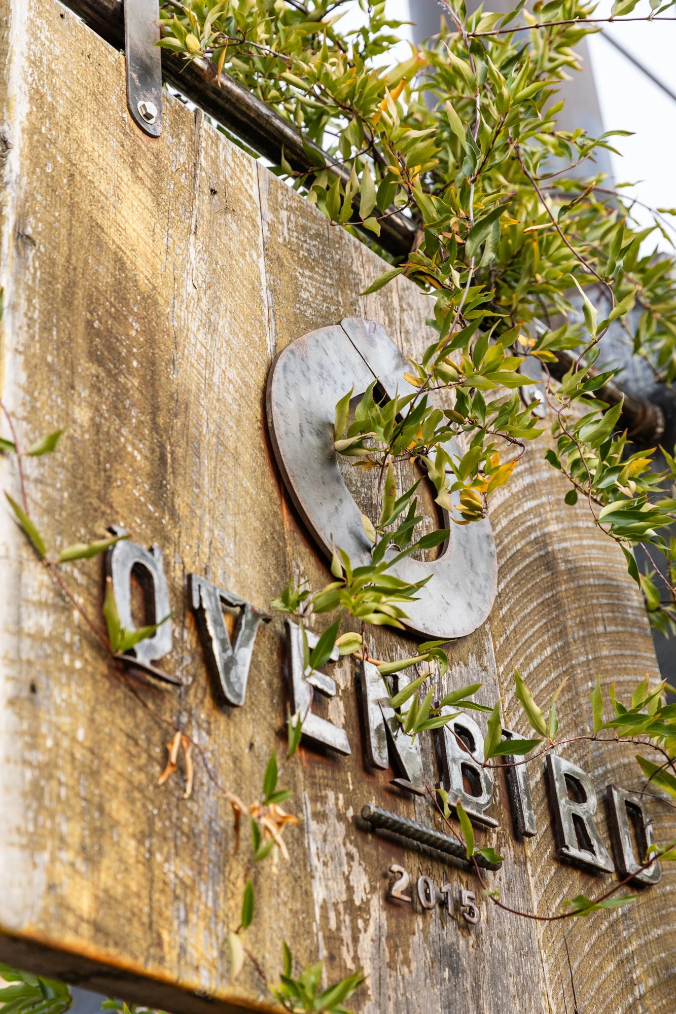 A rustic wooden sign with the words 'LOVE SHARE' in metal letters, surrounded by green vines and foliage, with the year 2015 inscribed on it. Ovenbird Restaurant in Birmingham, Alabama.