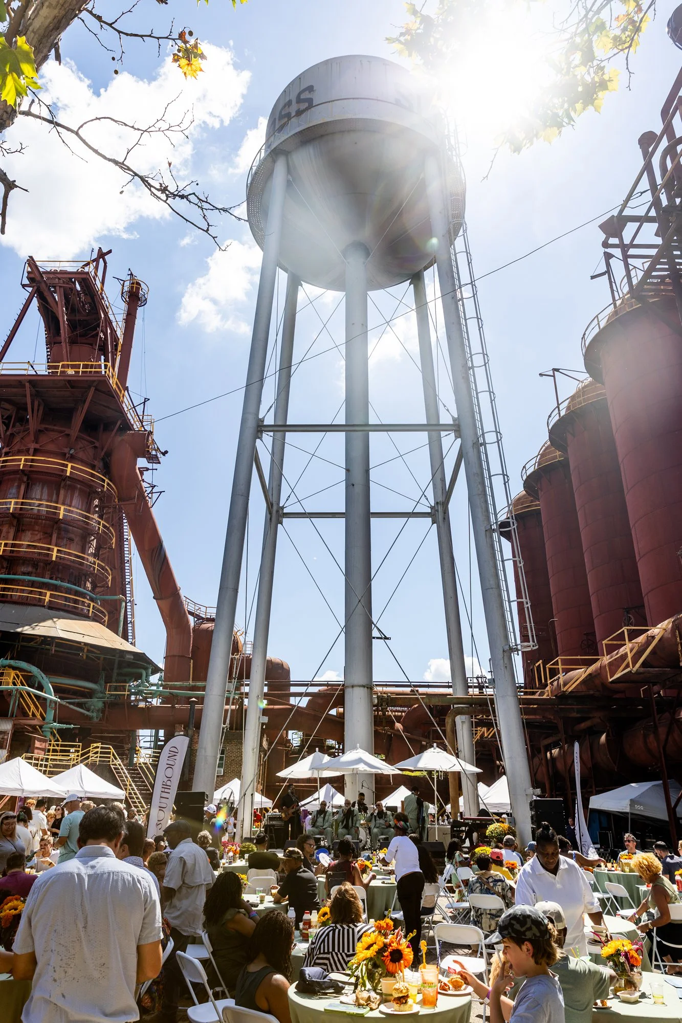 Outdoor event taking place in an industrial site with a tall water tower, surrounded by large rust-colored tanks and pipes. People are sitting at tables with floral arrangements and eating under umbrellas, while a band performs on a stage ahead. The 
