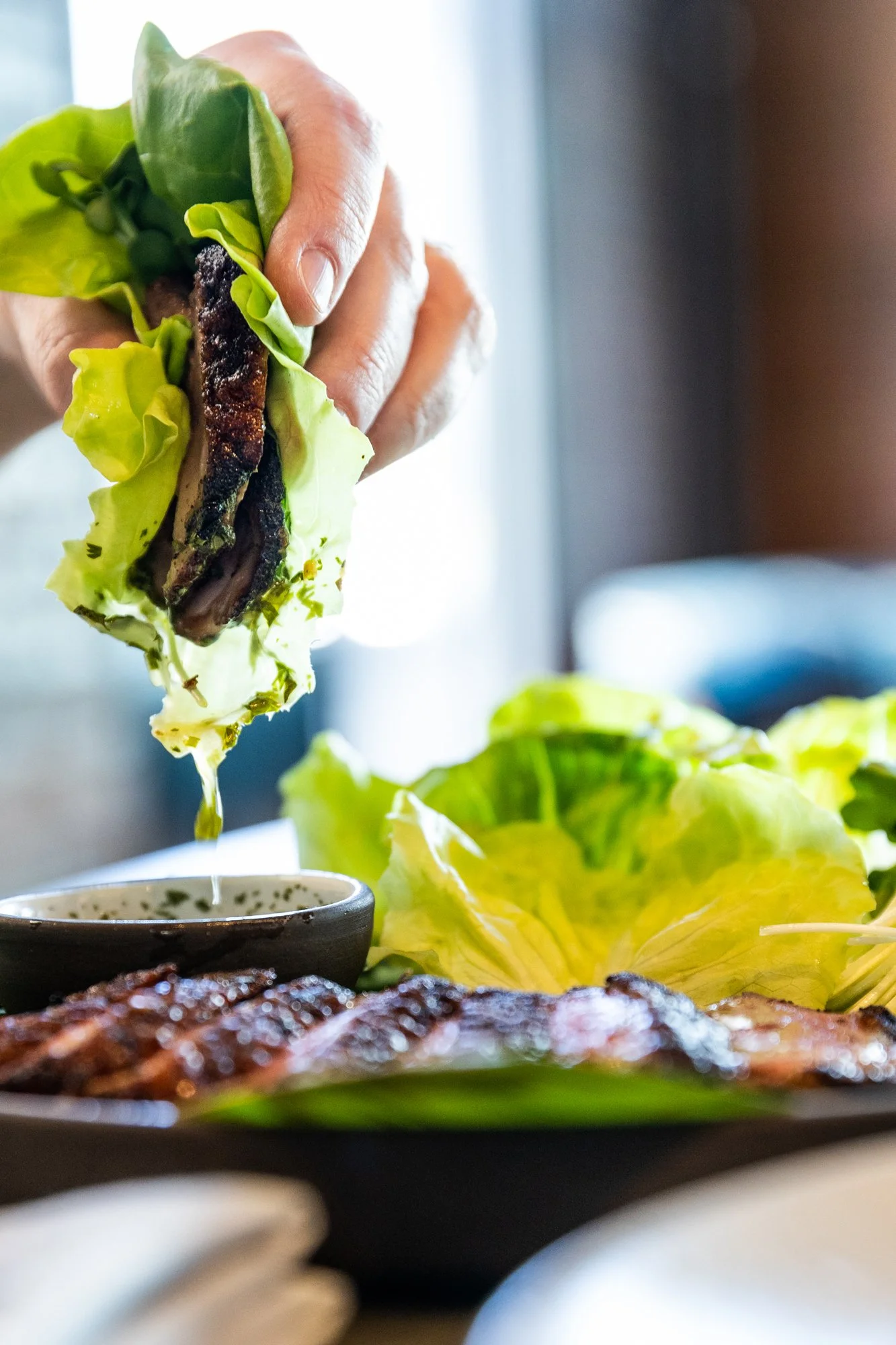 A person holding a piece of grilled meat wrapped in lettuce, drizzling sauce or oil onto a plate with grilled meat and fresh lettuce leaves.