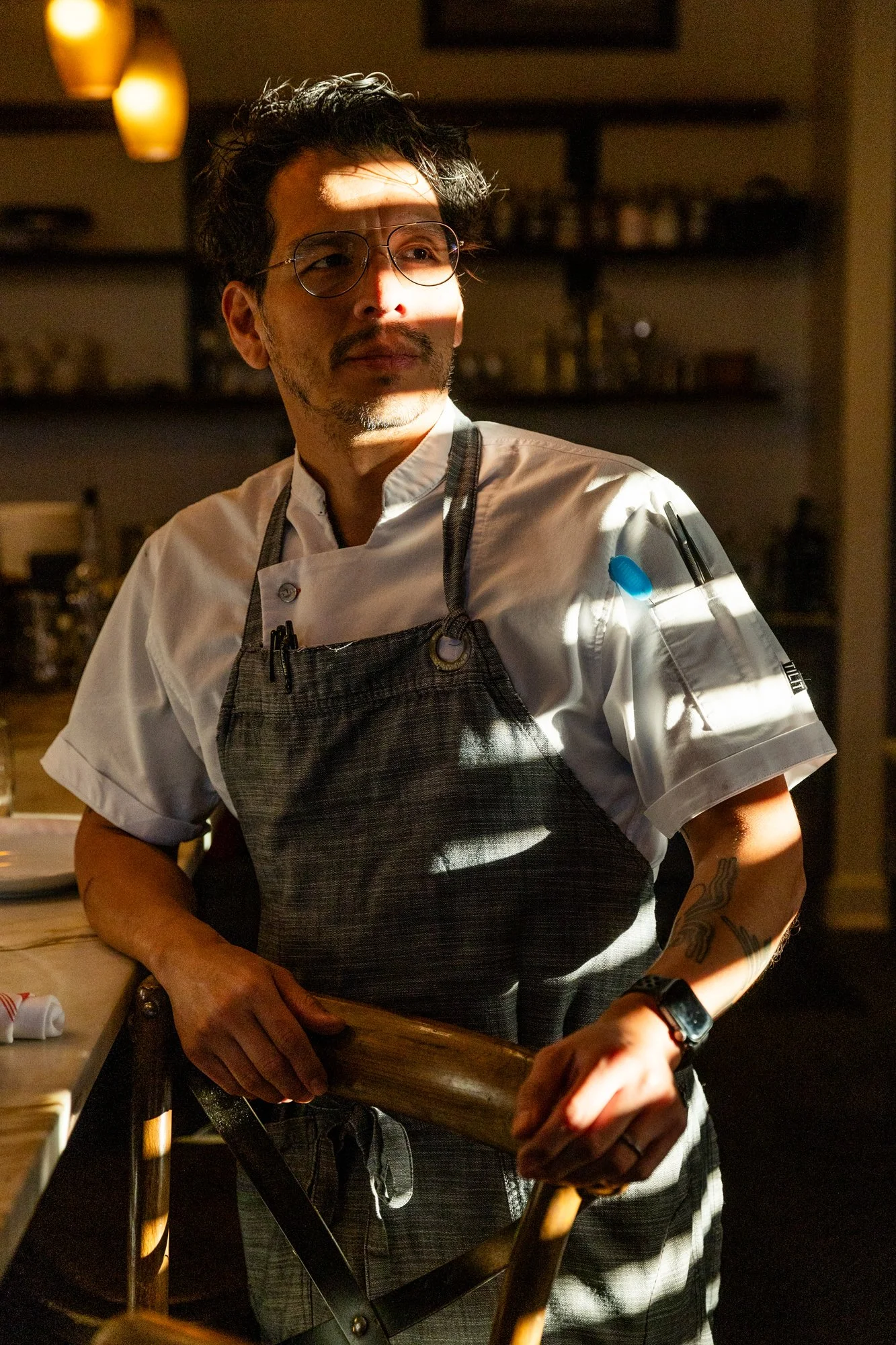 A male chef with glasses and a beard holds a wooden chair in a kitchen with warm lighting and shelves of jars and bottles behind him. Ovenbird Restaurant in Birmingham, Alabama.