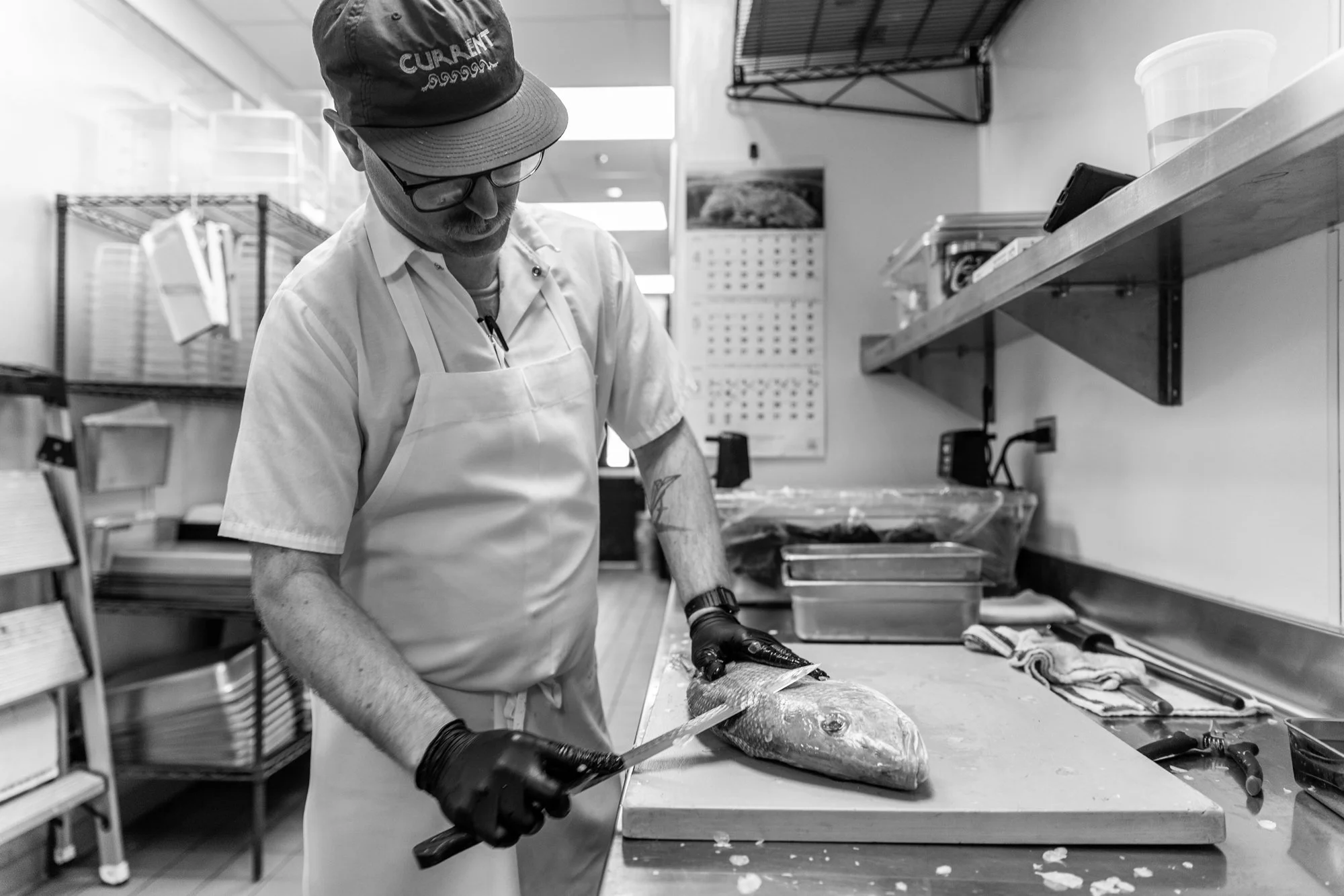 A chef wearing a apron, cap, and gloves is preparing a fish on a cutting board in a commercial kitchen.