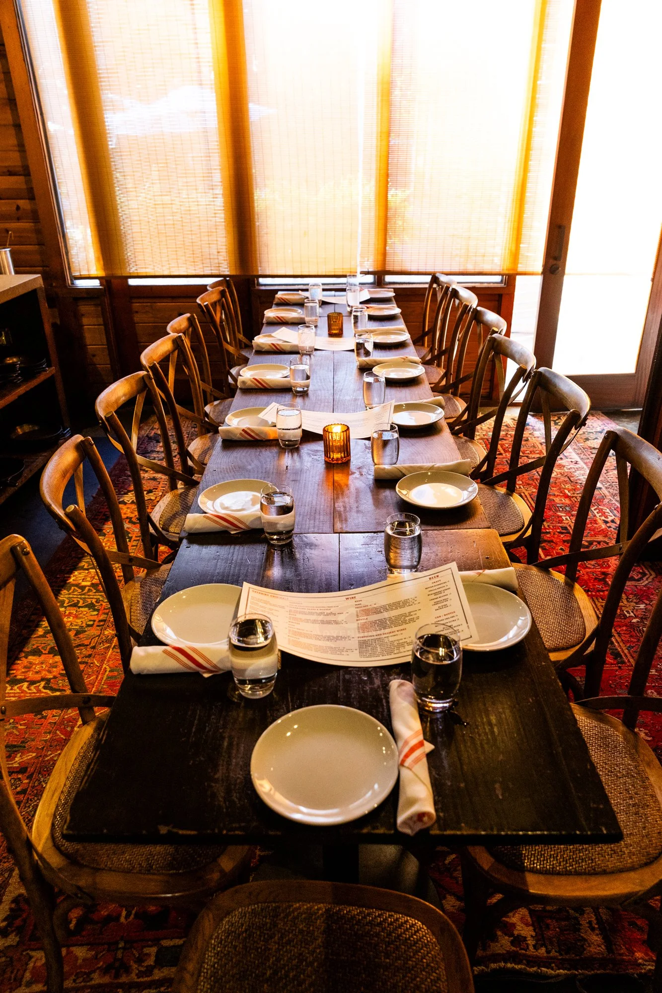 A long wooden dining table set for a meal with plates, napkins, glasses of water, and small candles, in a cozy restaurant with windows covered by orange blinds, wooden walls, and patterned carpet. Ovenbird Restaurant in Birmingham, Alabama.