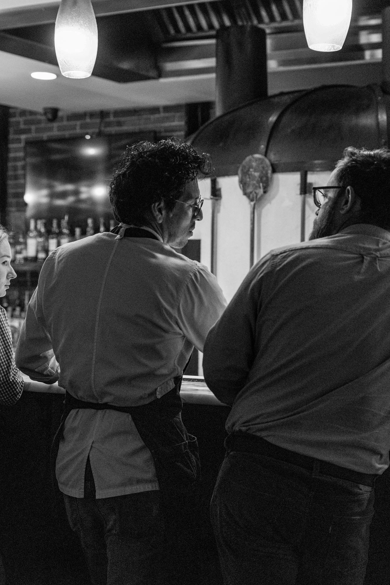 Two chefs in a restaurant kitchen, wearing aprons, working together at a counter with a pizza oven in the background. Ovenbird Restaurant in Birmingham, Alabama.