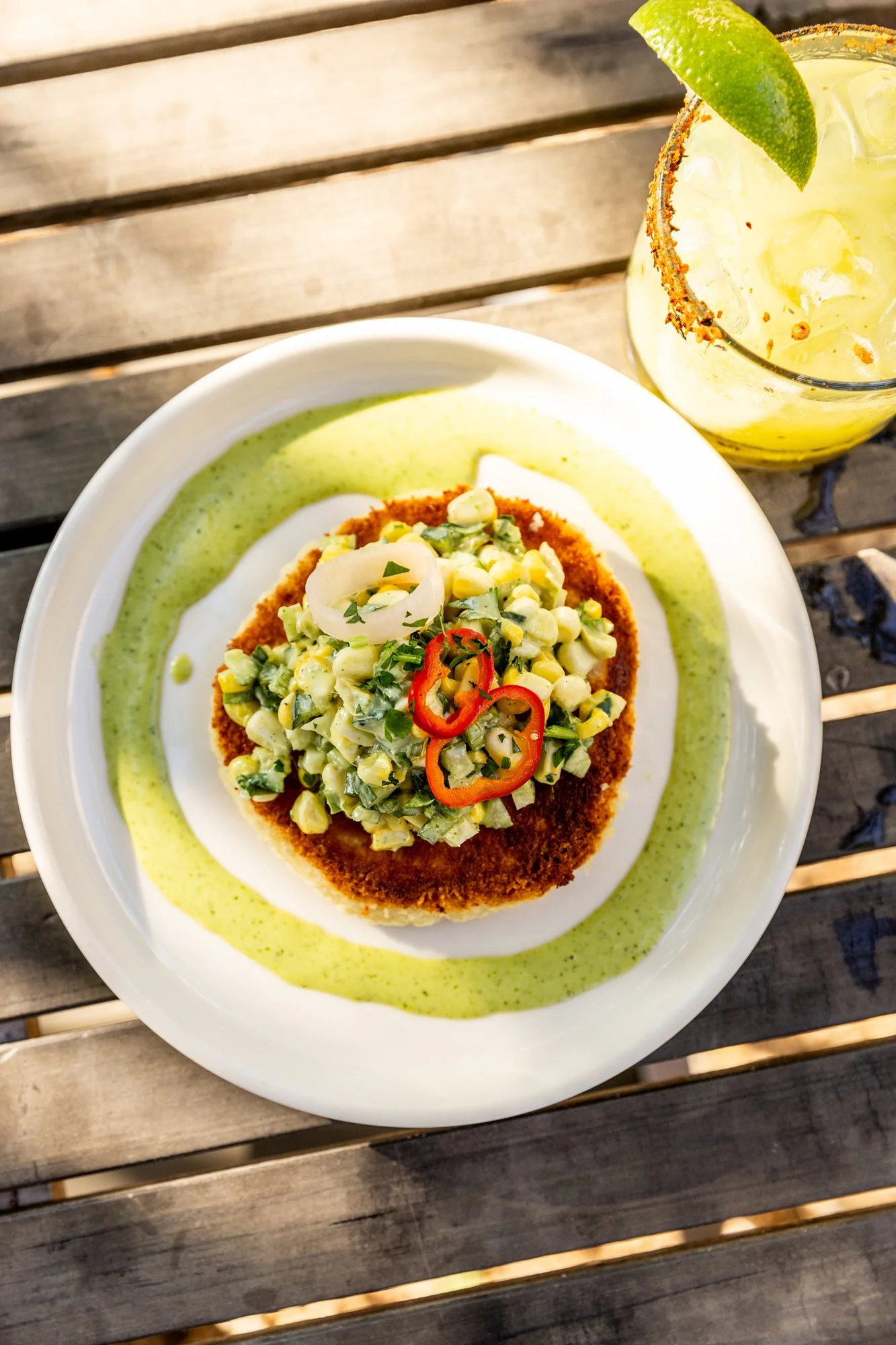 A plate with a breaded fried item topped with chopped vegetables and sliced chili peppers, accompanied by a green sauce and a lime wedge, with a glass of a yellow beverage garnished with a lime wedge in the background. Ovenbird Restaurant in Birmingh
