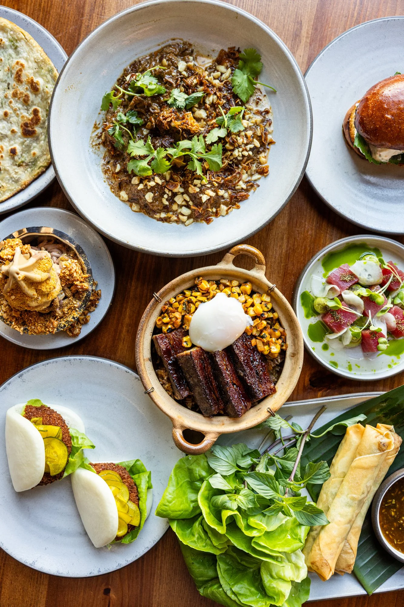 A variety of Asian dishes including braised beef, grilled ribs with a poached egg, spring rolls, a sandwich with pickles, and various vegetable dishes on a wooden table.
