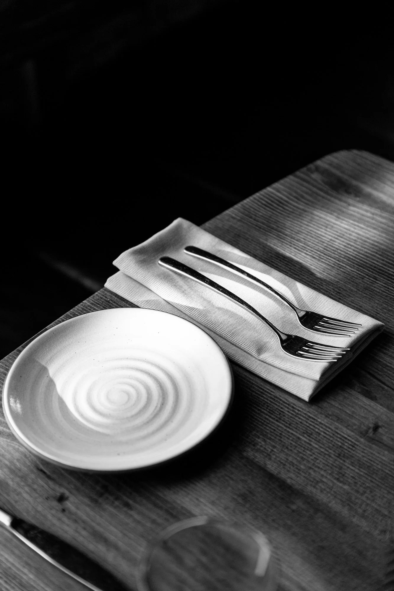 A black and white photo of a place setting with a small plate, a napkin, and two forks on a textured wooden table.