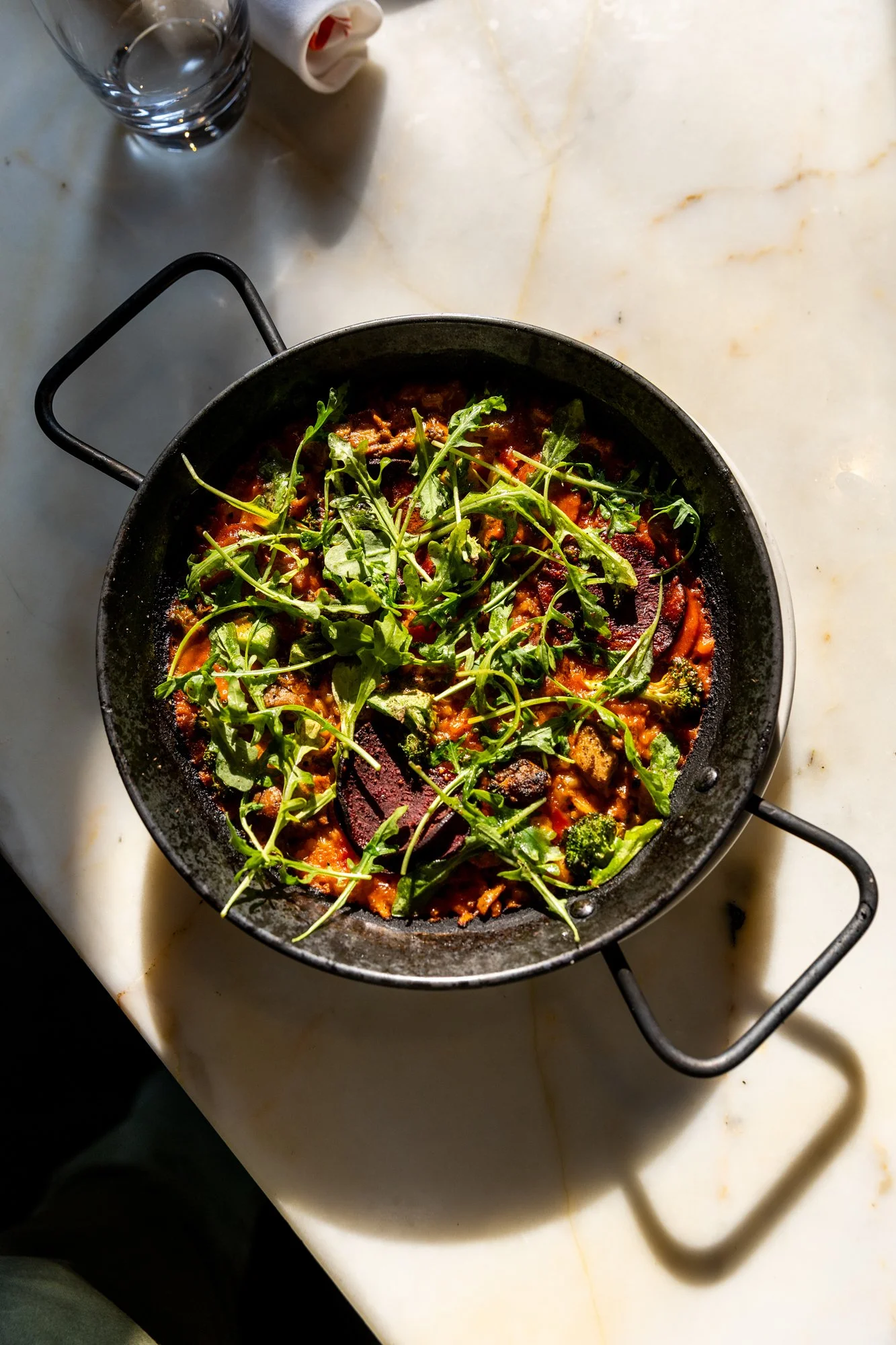A black skillet of cooked vegetable stew topped with fresh arugula on a white marble table. Ovenbird Restaurant in Birmingham, Alabama.
