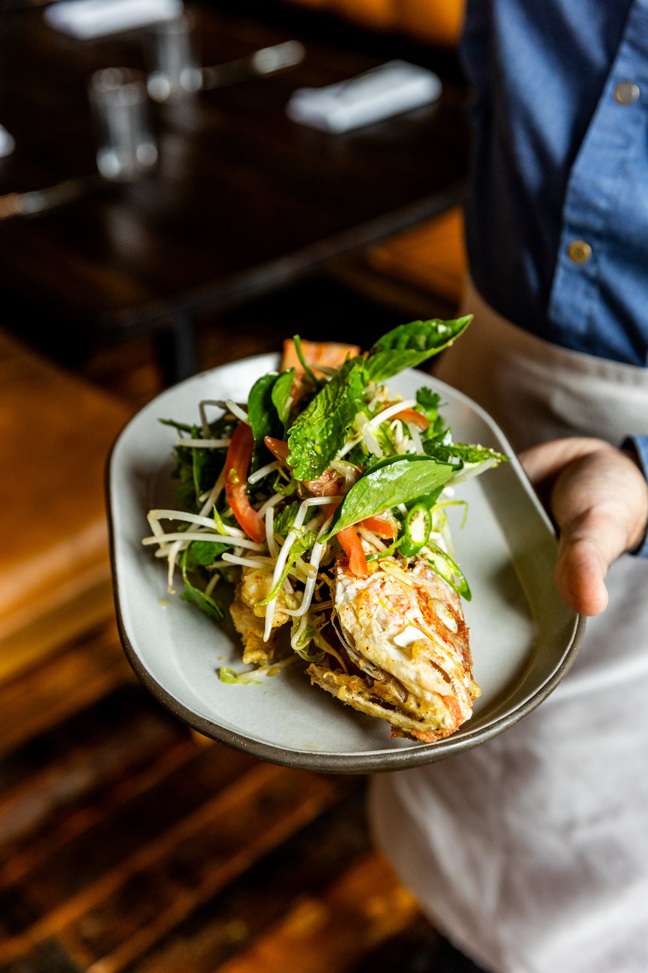 A person holding a plate with a seafood salad and vegetables. The person is wearing a blue shirt and beige apron. The background shows a dark wooden table.
