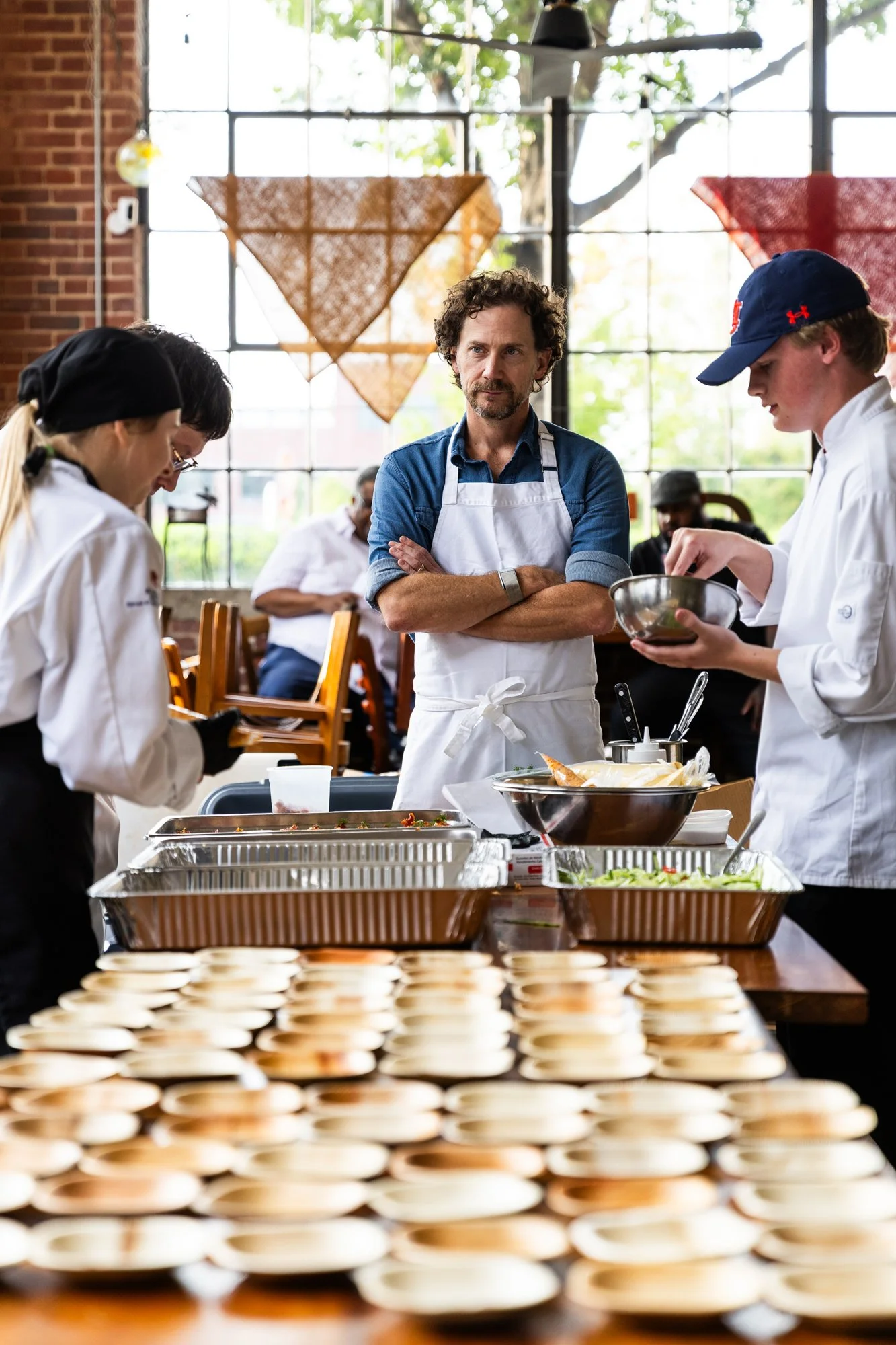 People preparing food in a restaurant kitchen with trays of plates on a table in the foreground. An instructor or chef observes as others cook. Southbound Food Festival in Birmingham, Alabama.