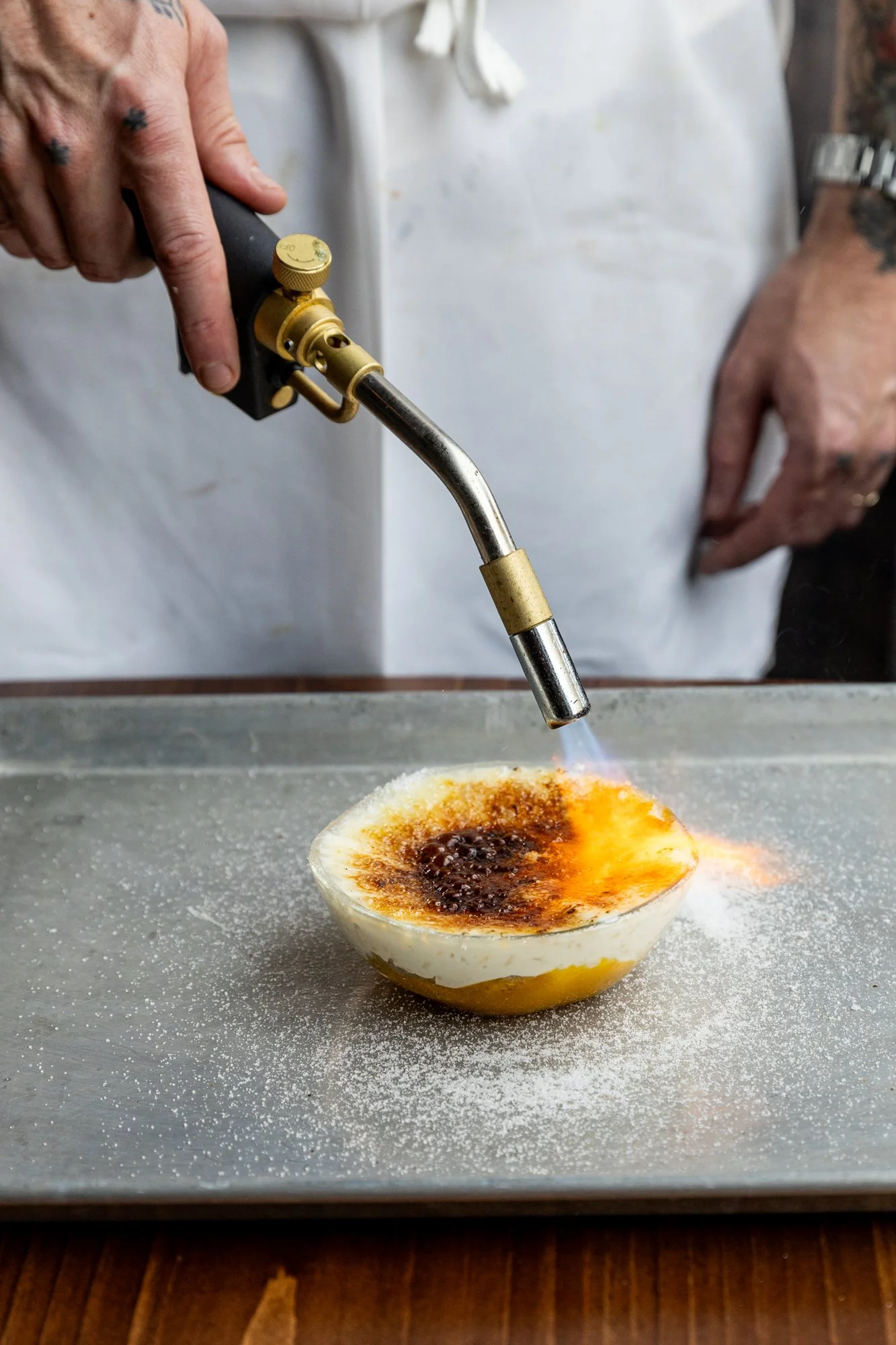 A person wearing a white apron uses a handheld culinary blowtorch to caramelize the top of a dessert in a bowl, with sugar sprinkled around on a metallic surface.