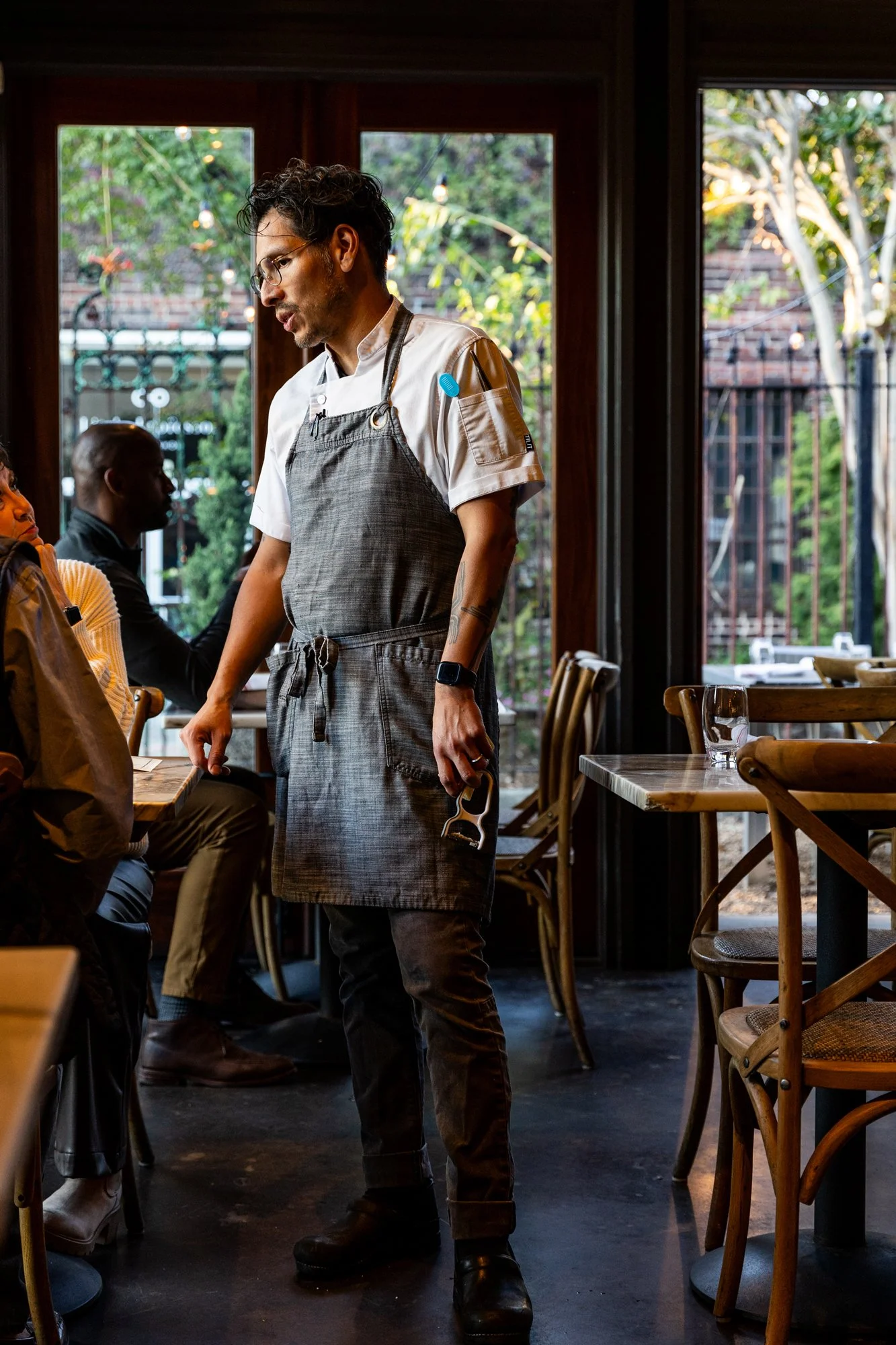 A male chef dressed in a gray apron and white shirt standing inside a restaurant, holding a corkscrew, engaging with a seated guest at a table. The restaurant has wooden furniture, large windows, and green outdoor scenery visible. Ovenbird Restaurant