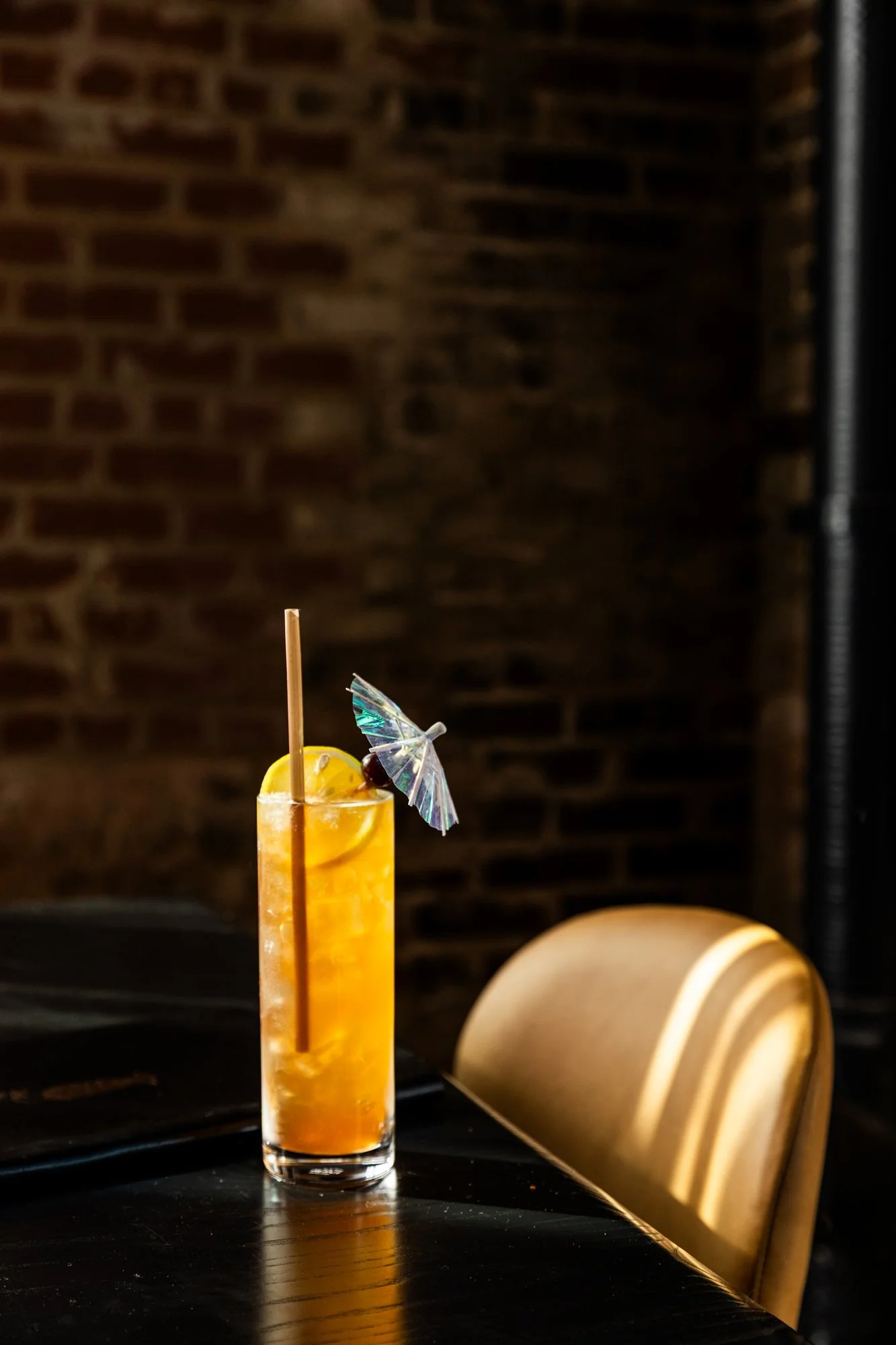 A tall glass of orange cocktail with a lemon slice garnish and a small decorative umbrella, sitting on a black table with a brick wall background. Birmingham, Alabama.