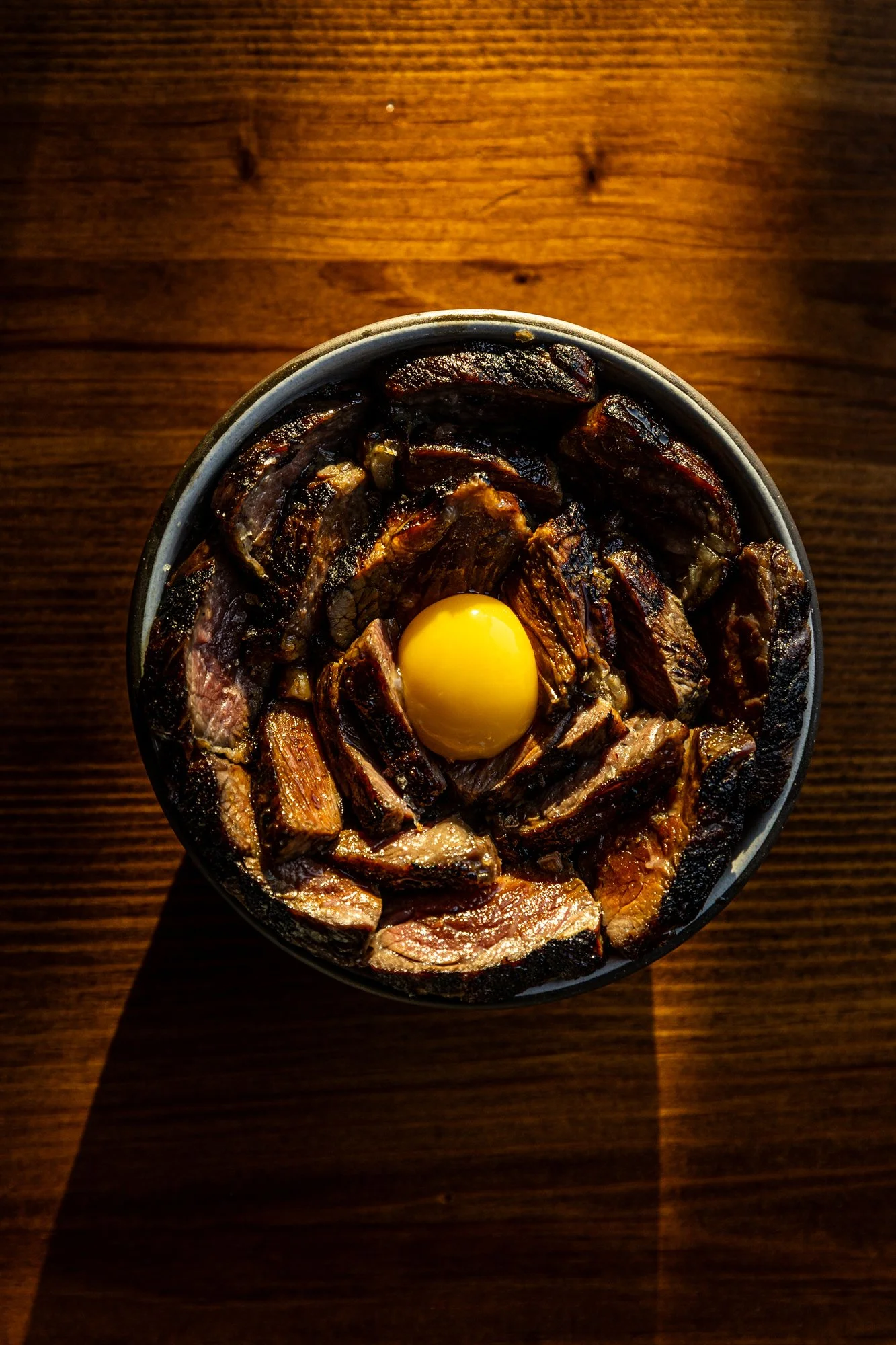 A bowl of cooked beef ribs with a raw egg yolk on top, placed on a wooden surface.