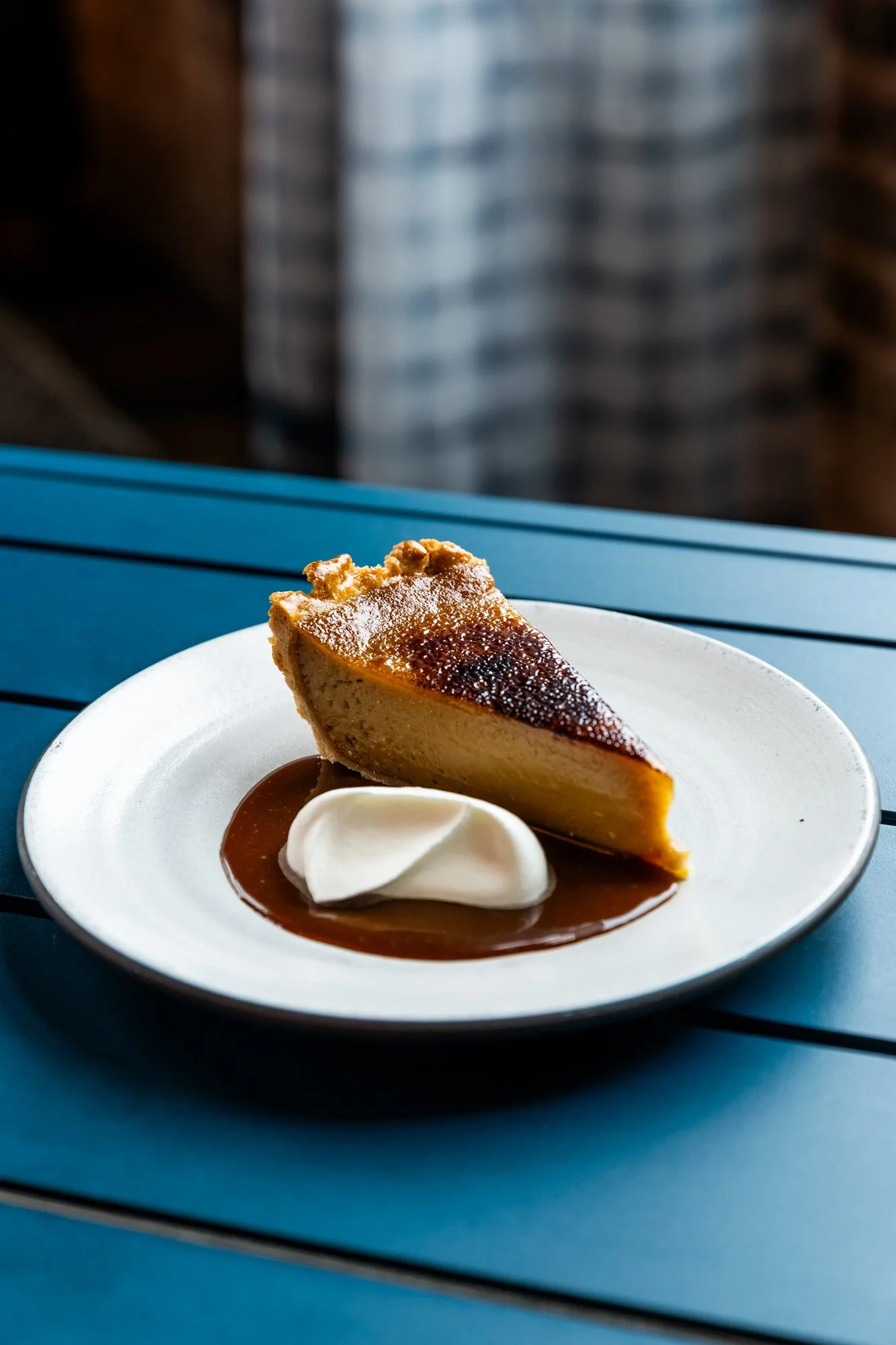 Slice of cheesecake on a white plate with dollop of whipped cream and chocolate sauce, on a blue table.