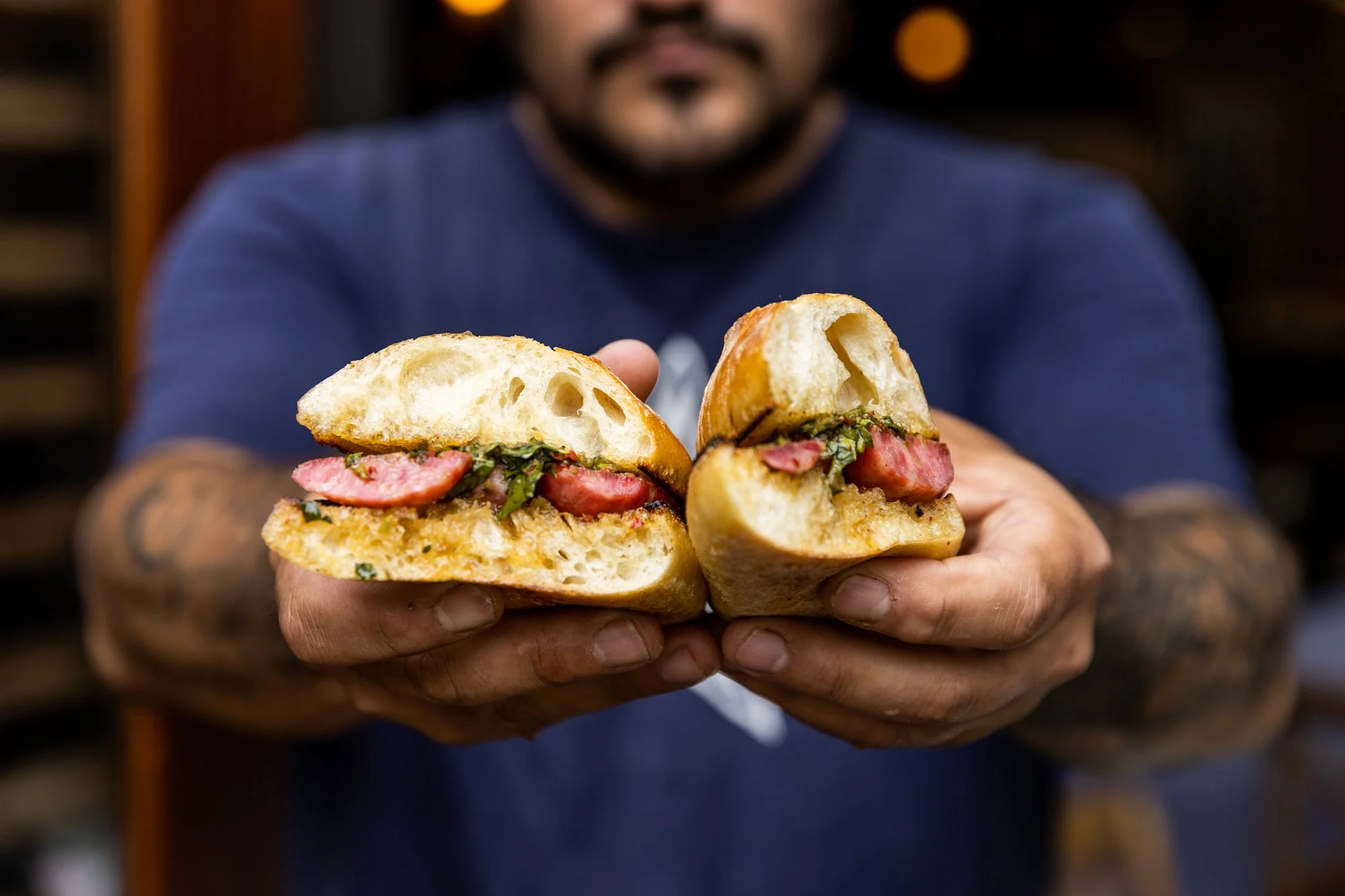 A man holding a split sandwich with visible bacon, greens, and bread, with a blurred background. Ovenbird Restaurant in Birmingham, Alabama.