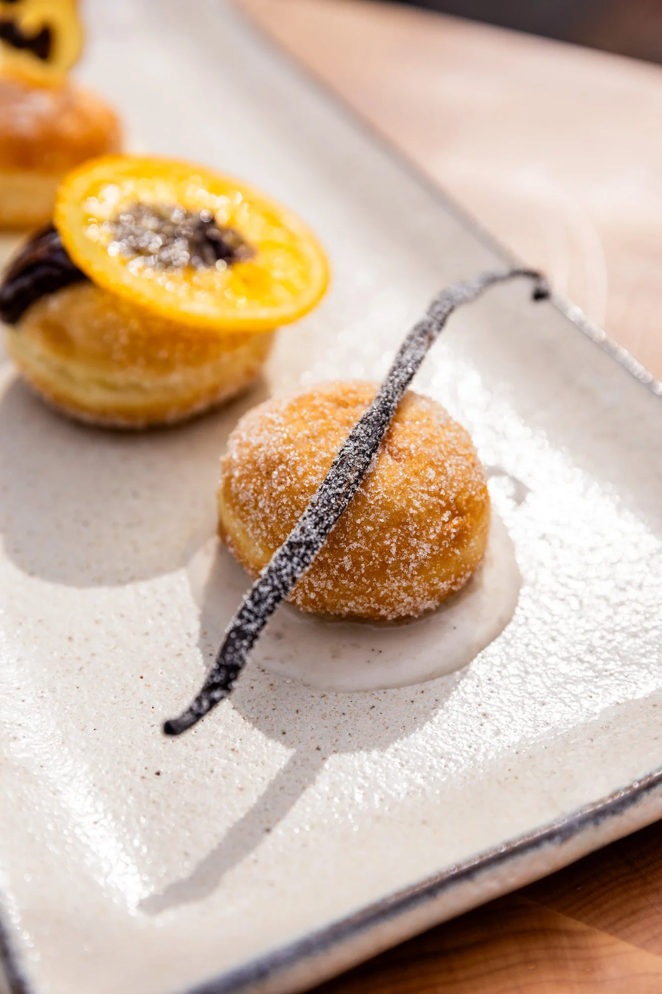 A dessert featuring a round, golden fried ball dusted with powdered sugar, placed on a white sauce and garnished with a thin chocolate strip. In the background, there is a halved passion fruit with yellow pulp and black seeds. Hot and Hot Fish Club R