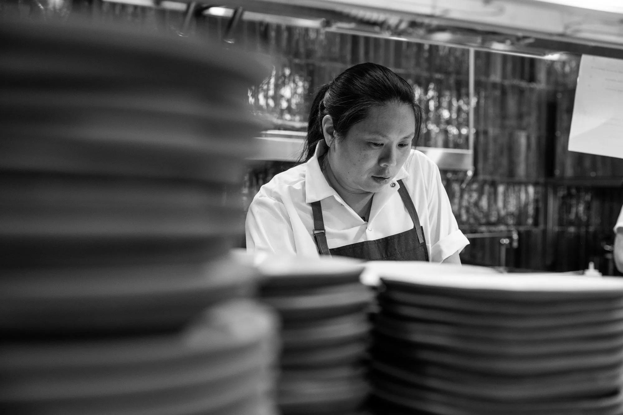 A woman in a white shirt and apron working in a kitchen, surrounded by stacks of plates.