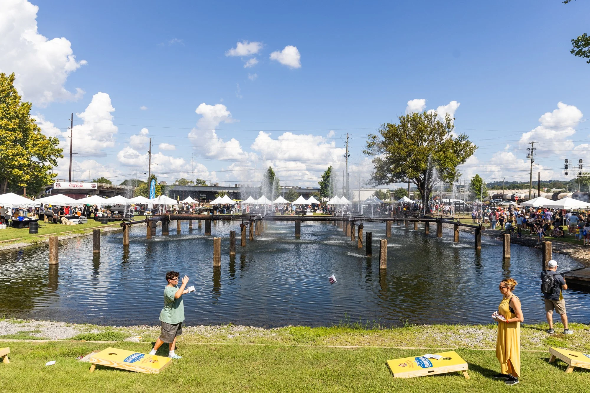 Outdoor fair scene with tents, crowd, and a small pond with fountains under a blue sky with scattered clouds. Southbound Food Festival in Birmingham, Alabama.