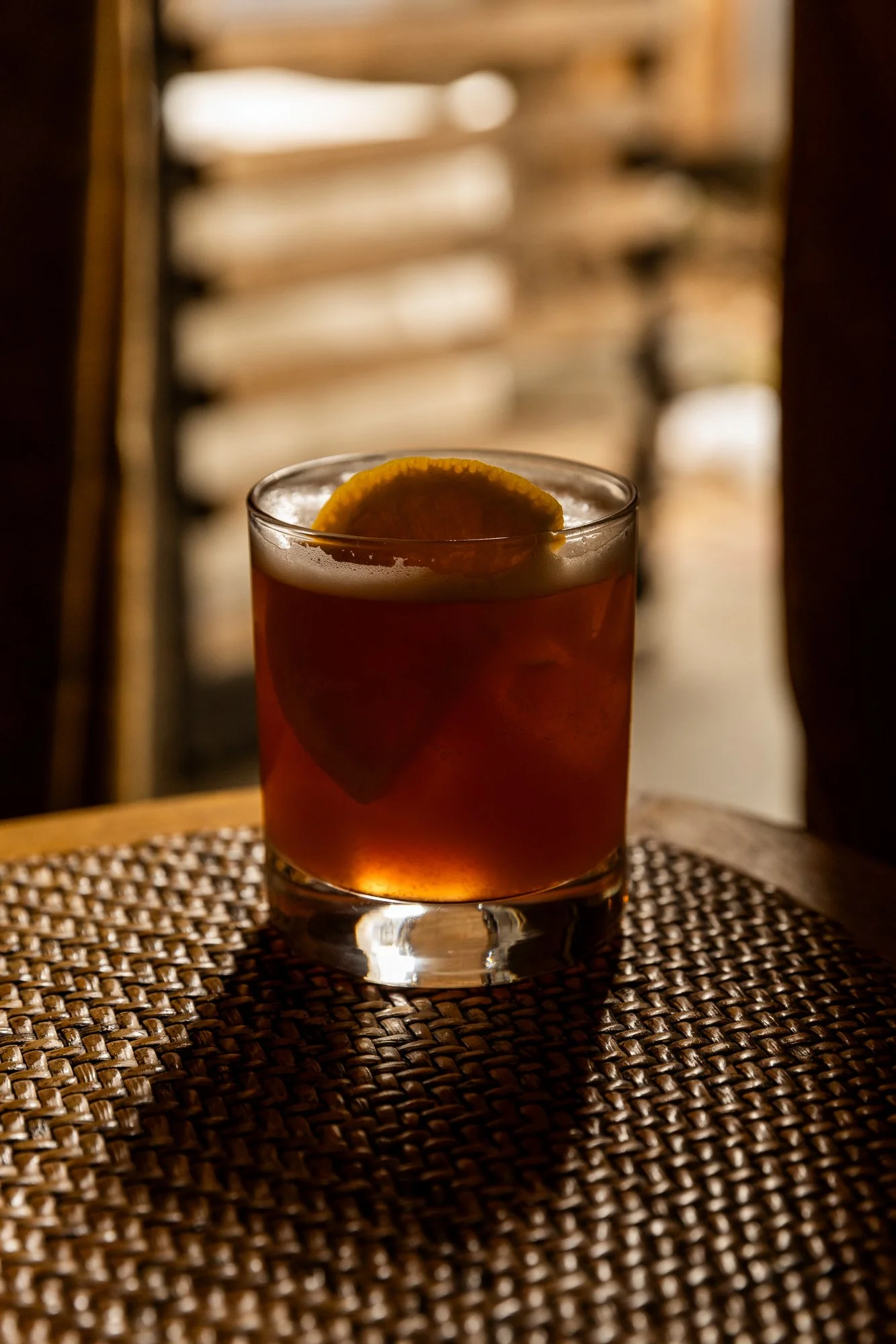 A glass of a dark cocktail garnished with a lemon slice, placed on a woven placemat with a blurred background of wooden slats. Ovenbird Restaurant in Birmingham, Alabama.