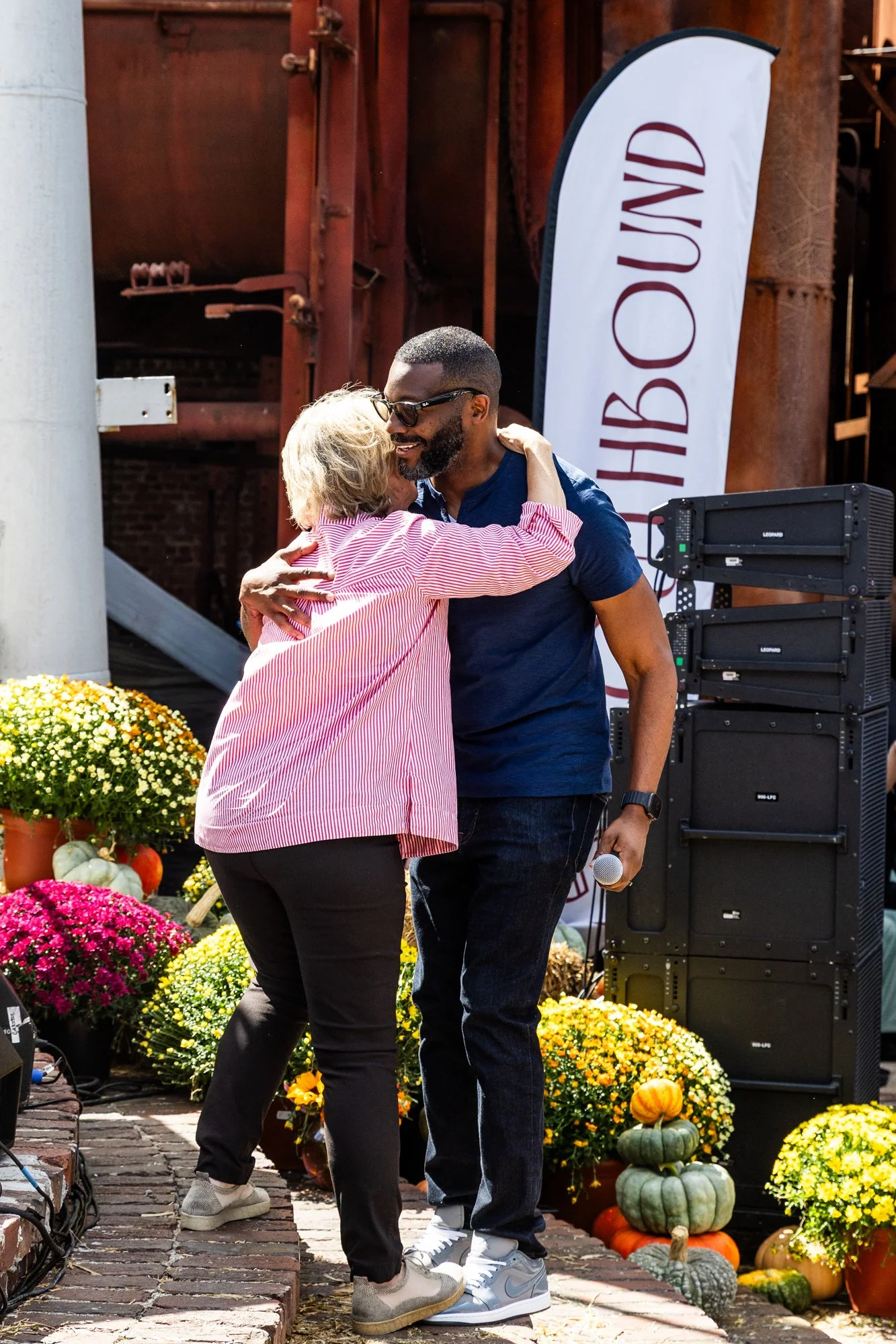 A man and an older woman hug on an outdoor stage decorated with pumpkins and flowers. The man is holding a microphone and wearing sunglasses, while the woman has blond hair and is dressed in a red and white striped shirt. Southbound Food Festival in 