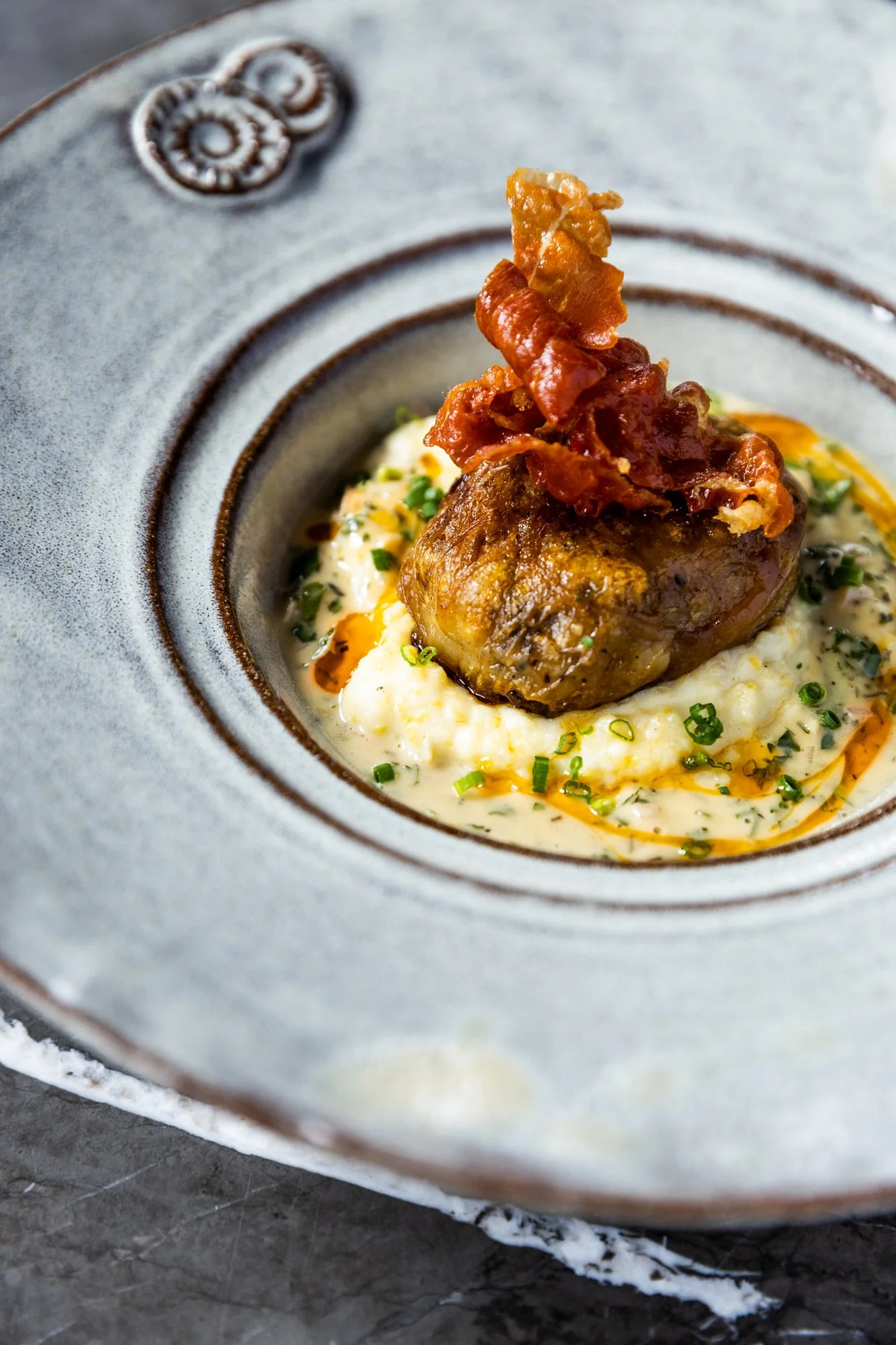A plated dish featuring a piece of roasted meat topped with crispy bacon, served on a bed of mashed potatoes with garnished green herbs and a drizzle of sauce, all presented on a rustic gray plate. Hot and Hot Fish Club Restaurant in Birmingham