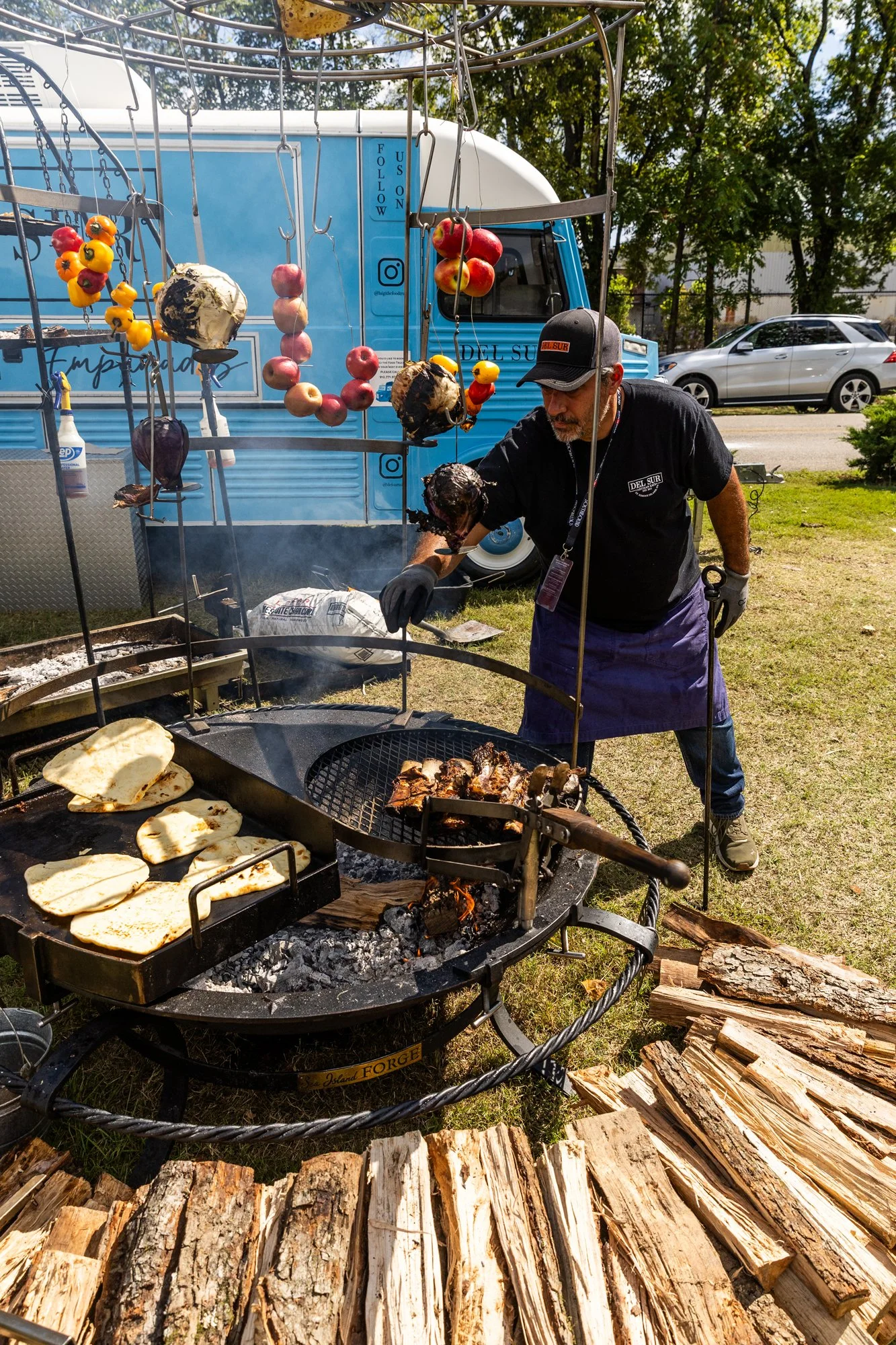 A man wearing a black shirt, purple apron, gray cap, and gloves is grilling meats and flatbreads over an open fire pit outdoors. There are wooden logs on the ground nearby and a food truck in the background with trees around. Southbound Food Festival