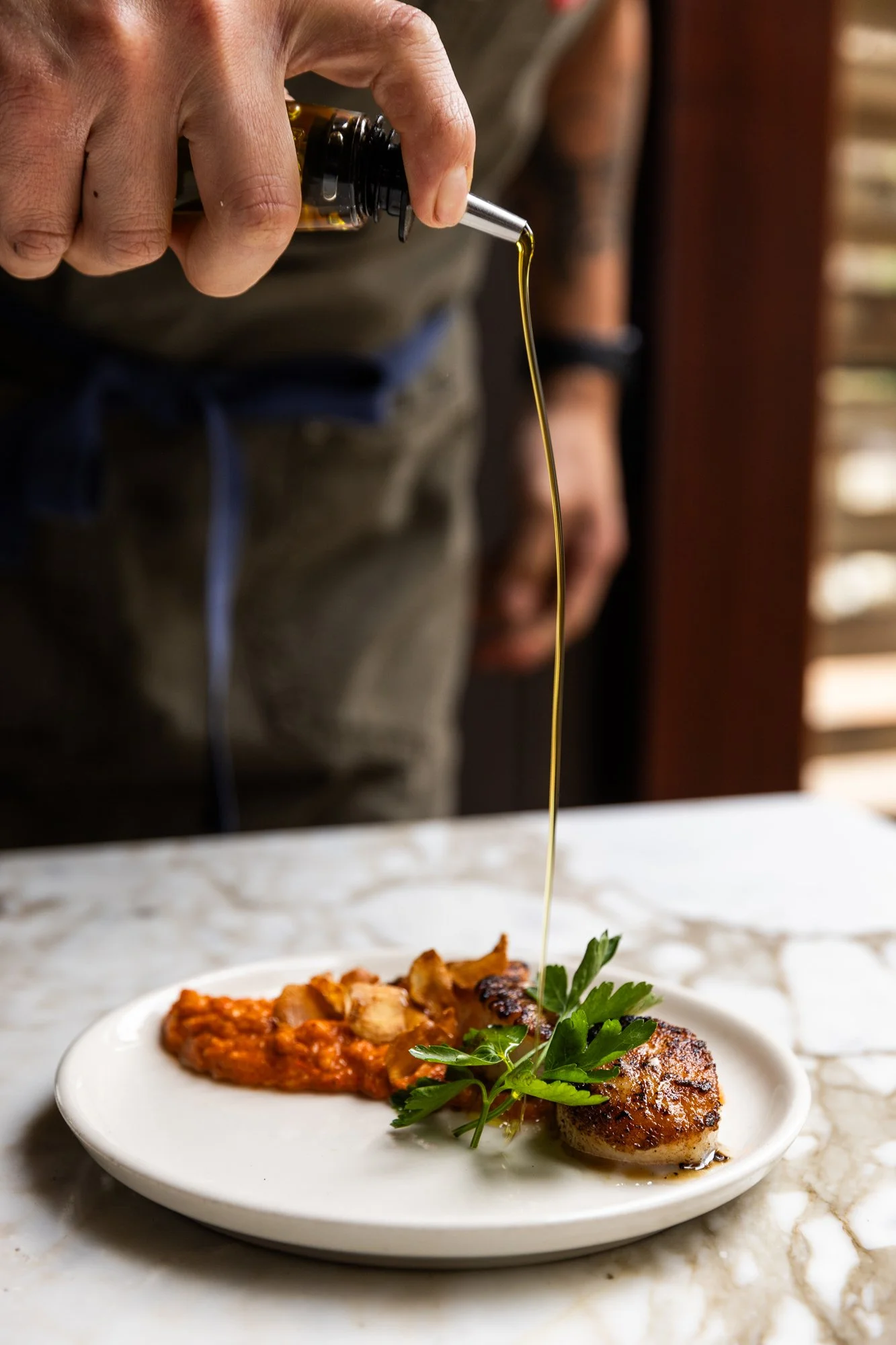Chef drizzling olive oil over a plated dish with meat, sauce, some greenery, and a side of vegetables on a white plate. Ovenbird Restaurant in Birmingham, Alabama.