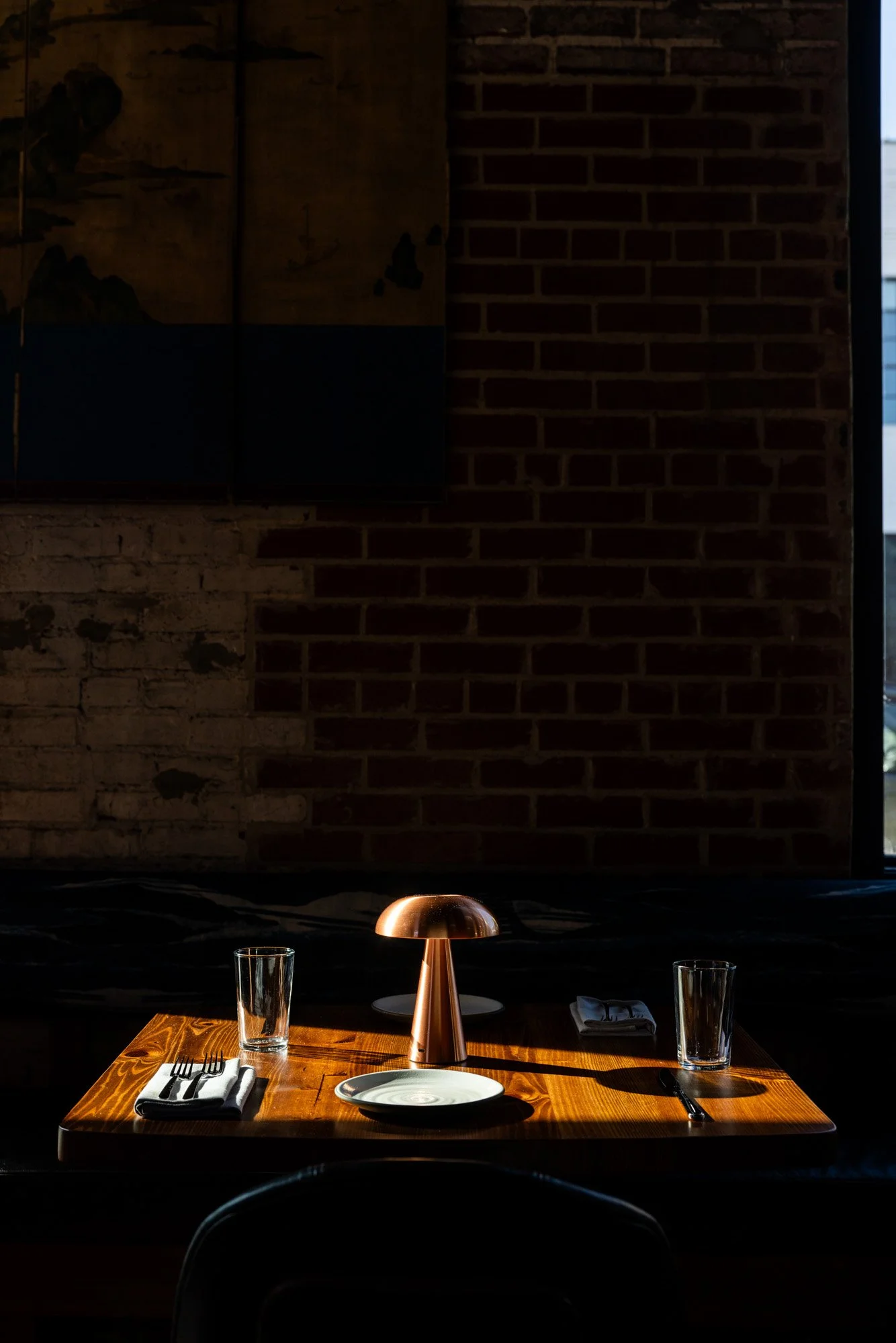 A restaurant table set with three glasses, two napkins, a fork, a knife, a white plate, and a small copper table lamp on a wooden table near a window, with a brick wall in the background.