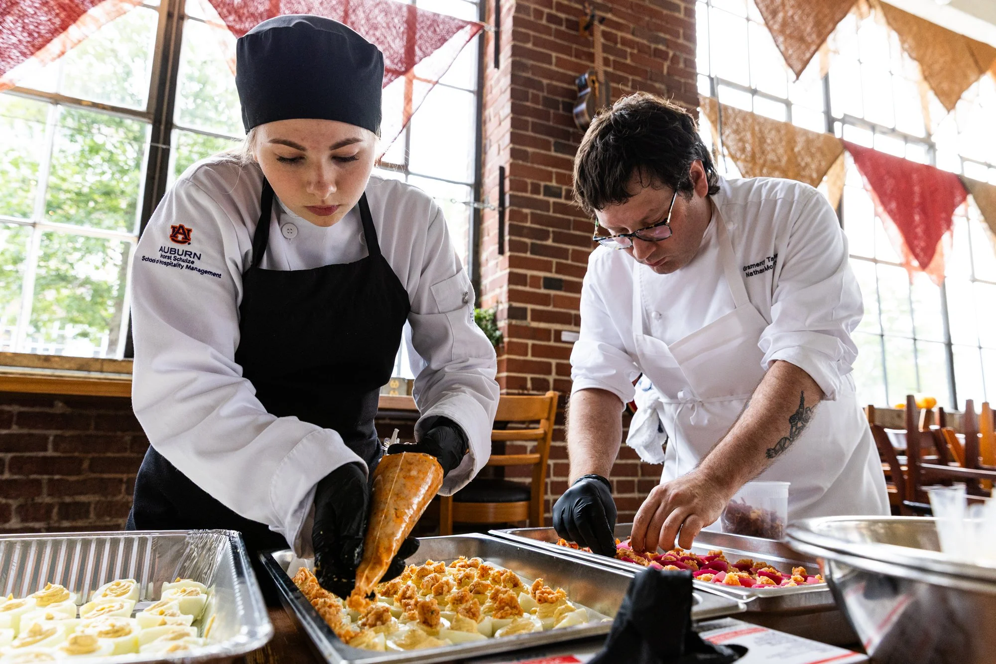 Two chefs in a kitchen preparing food, one female and one male, wearing white chef jackets and black gloves, with the female piping icing and the male arranging toppings on desserts. Southbound Food Festival in Birmingham, Alabama.
