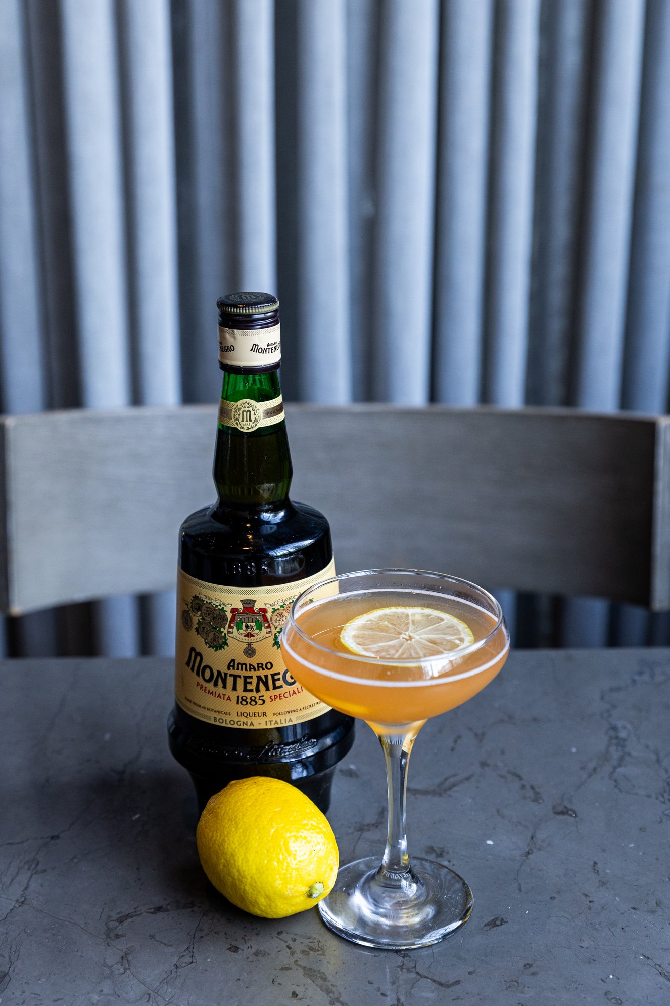 A cocktail in a coupe glass with lemon slice, a lemon, a bottle of Montenegro amaro, and a grey table with a wooden chair and a background of vertical metal panels. Hot and Hot Fish Club Restaurant in Birmingham, Alabama.