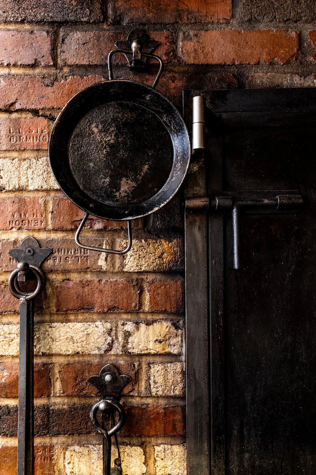 A black cast iron skillet hanging on a hook, mounted on a brick wall alongside other tools. Ovenbird Restaurant in Birmingham, Alabama.