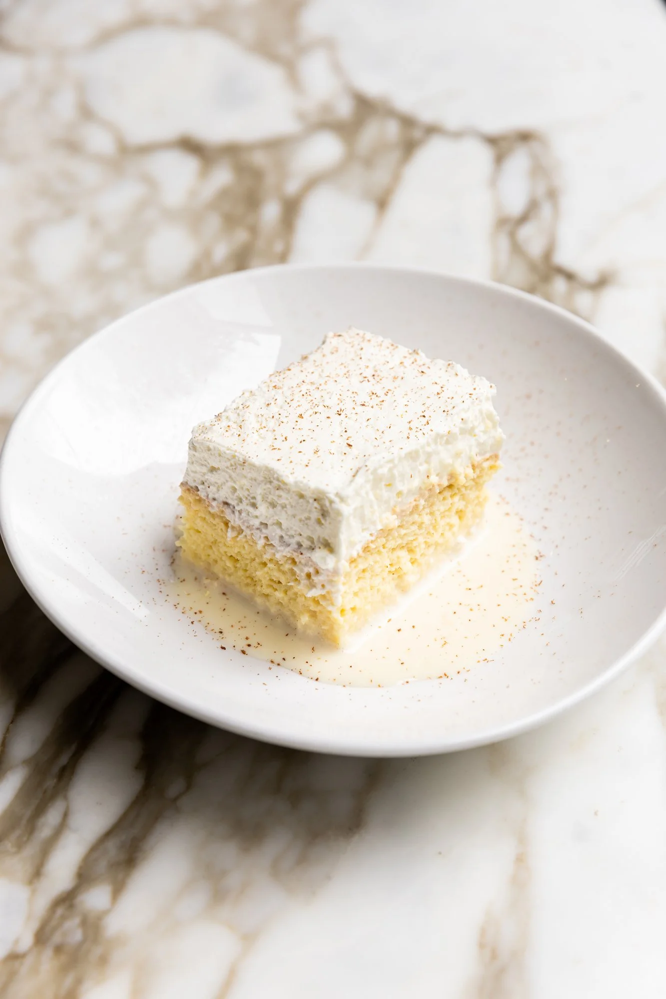 A square piece of vanilla cake with white frosting and a dusting of cinnamon on top, served on a white plate. Ovenbird Restaurant in Birmingham, Alabama.