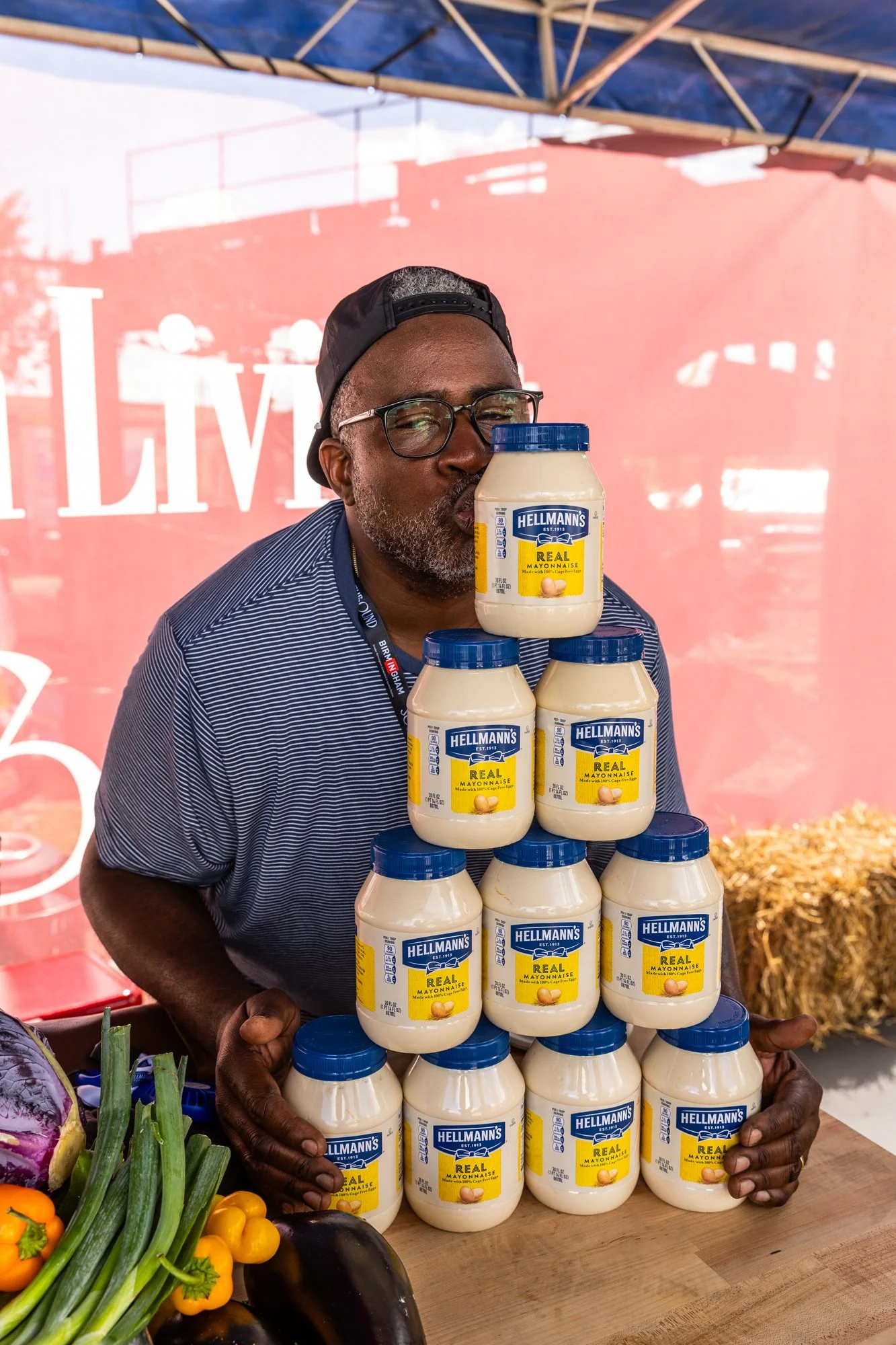 A man with glasses and a black cap displays a pyramid of Hellmann's mayonnaise jars at an outdoor event, with produce visible at the bottom left and a red banner in the background. Southbound Food Festival in Birmingham, Alabama.