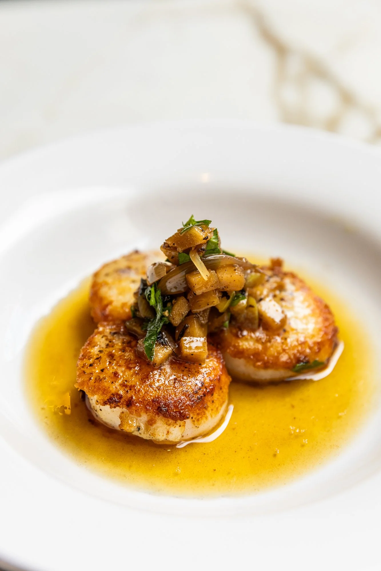 Seared scallops topped with a mushroom and green onion mixture, served in a yellow sauce on a white plate. Ovenbird Restaurant in Birmingham, Alabama.