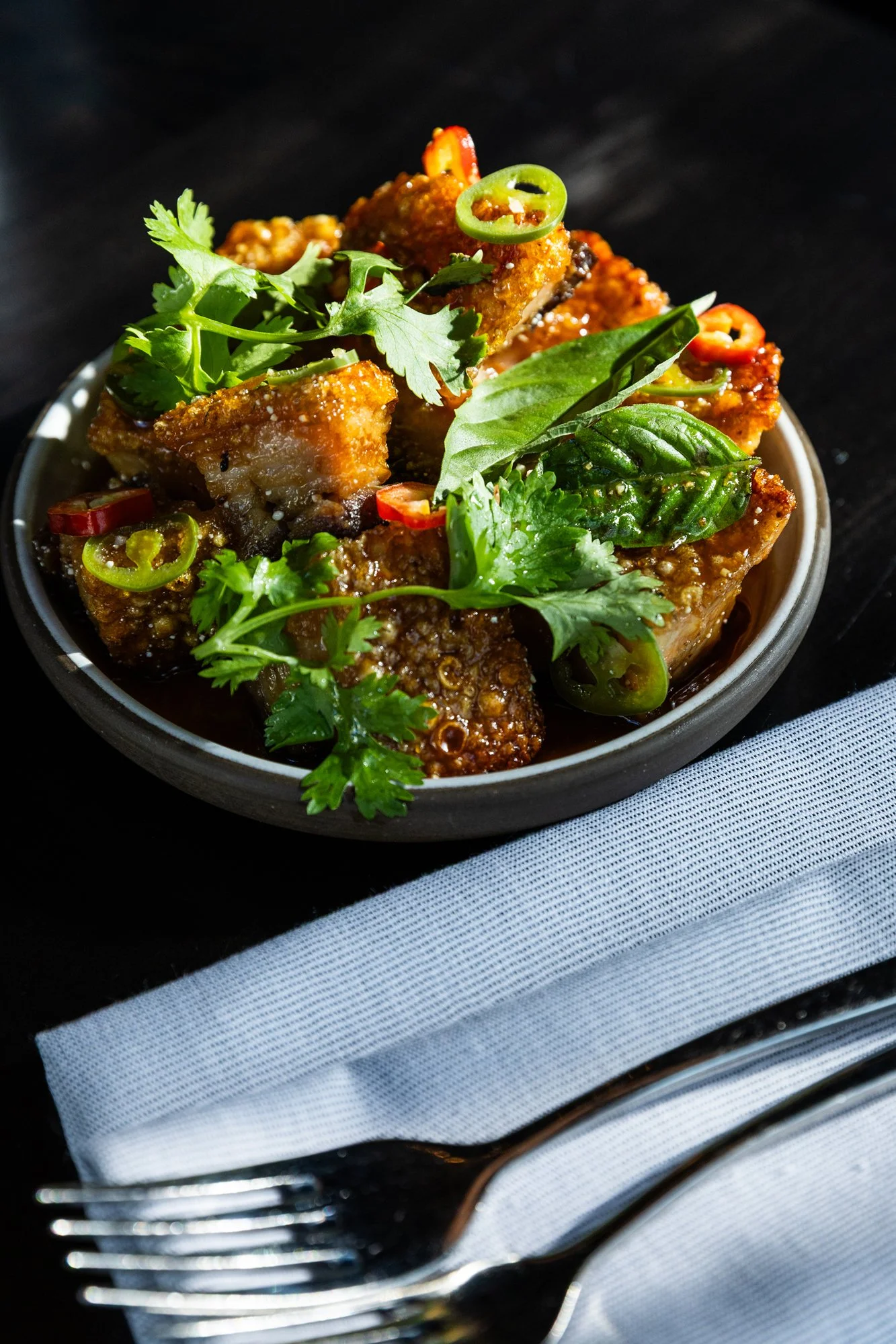 Close-up of a bowl of fried tofu garnished with cilantro, green and red chili slices, and fresh herbs, placed on a dark surface with a white napkin and silver fork nearby.