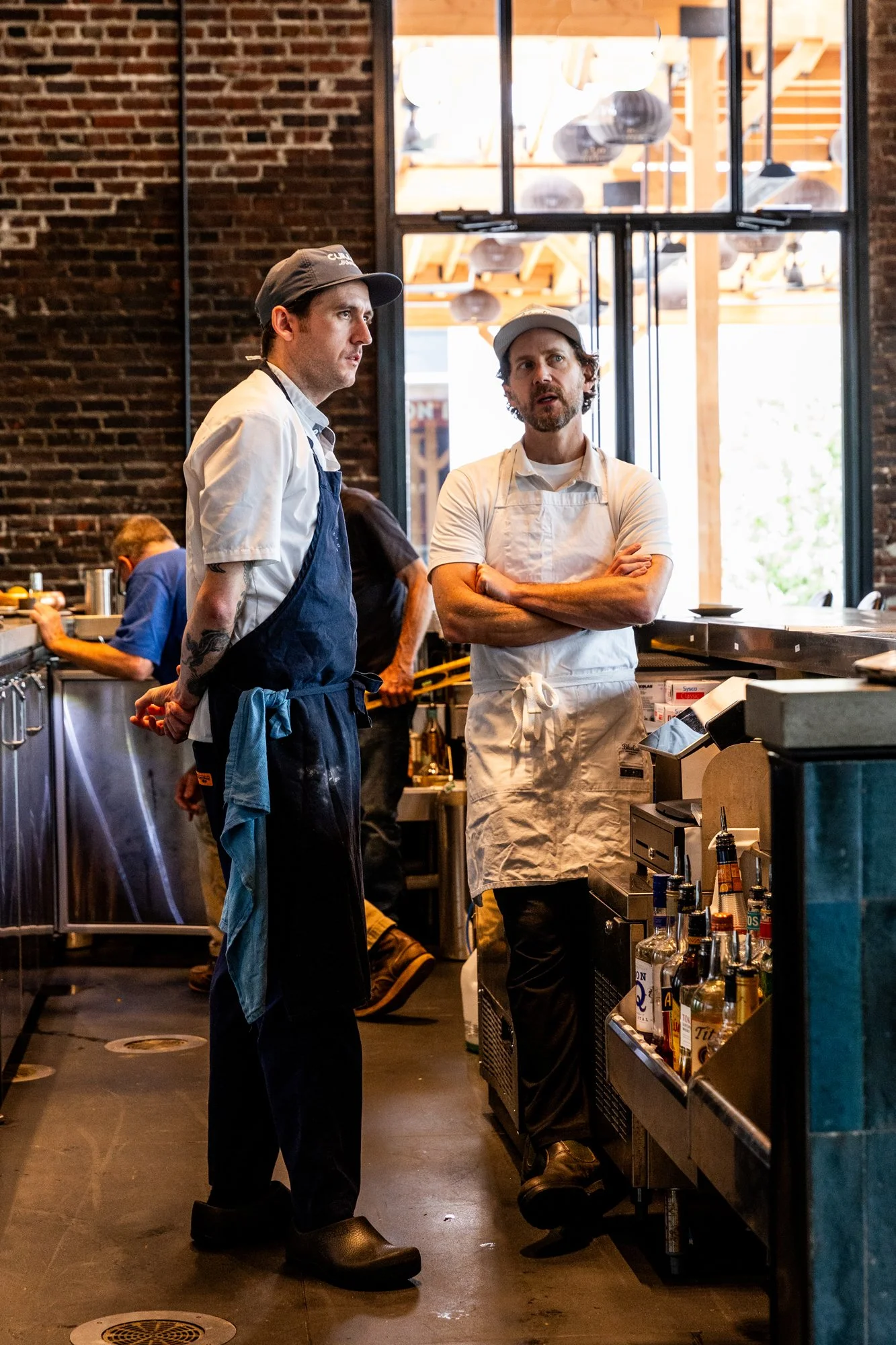 Two male chefs with aprons and hats standing in a restaurant kitchen, engaging in conversation. One has arms crossed, and the other has hands behind his back. Kitchen supplies and bottles are visible nearby, with a brick and glass window background a