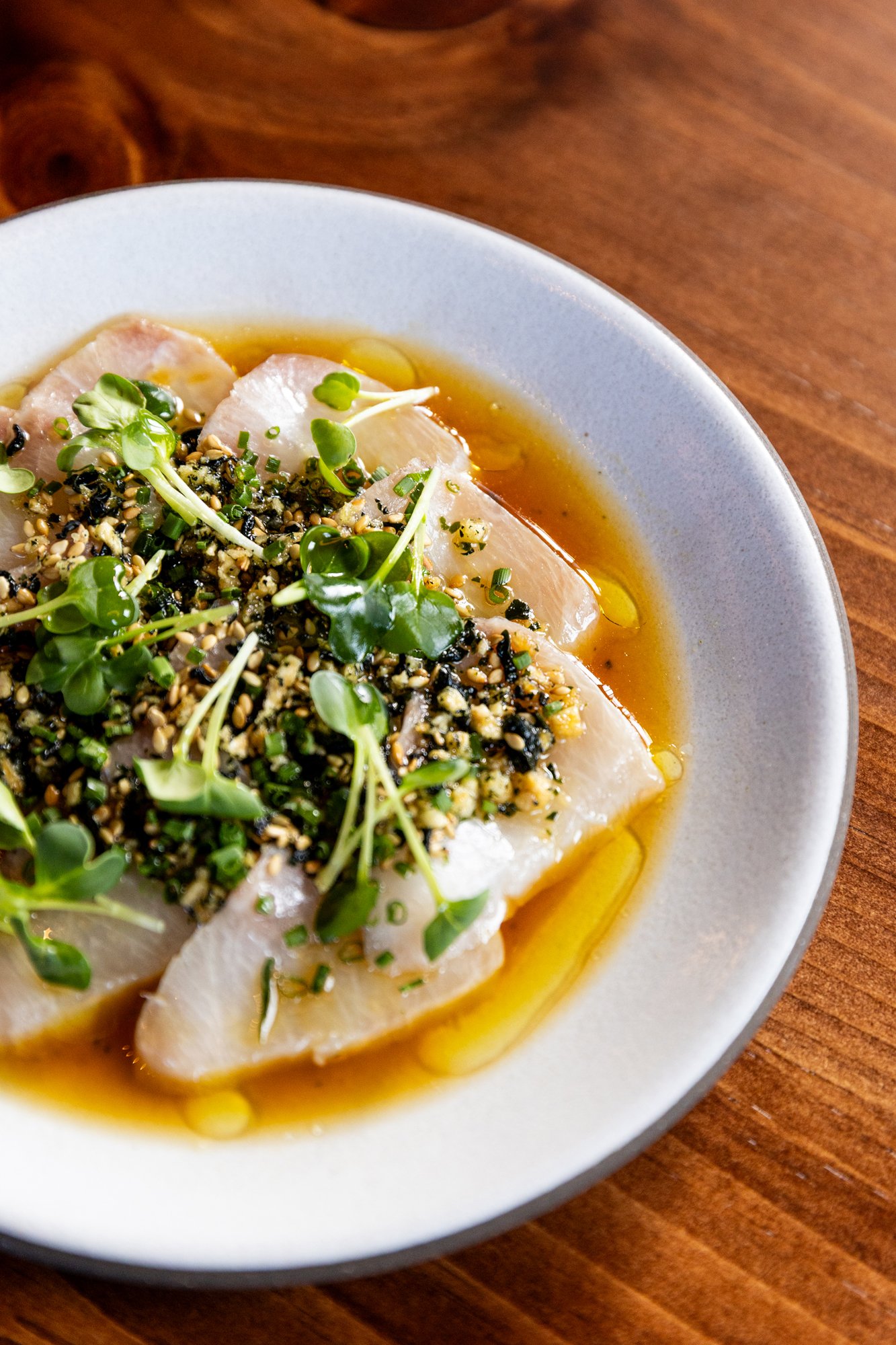 Slices of fish topped with chopped herbs and microgreens, drizzled with oil and sauce, served on a white bowl over a wooden surface.