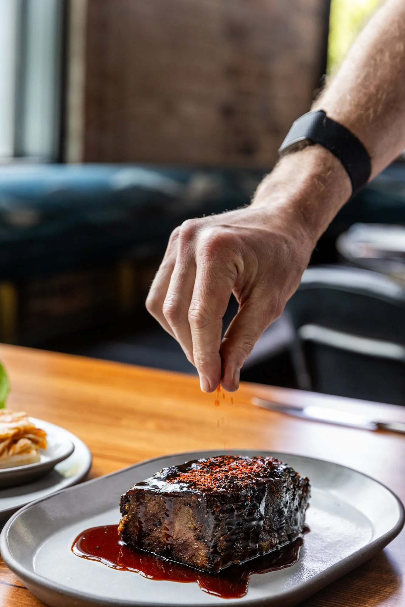 A person's hand sprinkling orange spices over a cooked meat dish with dark sauce, on a gray plate, on a wooden table.
