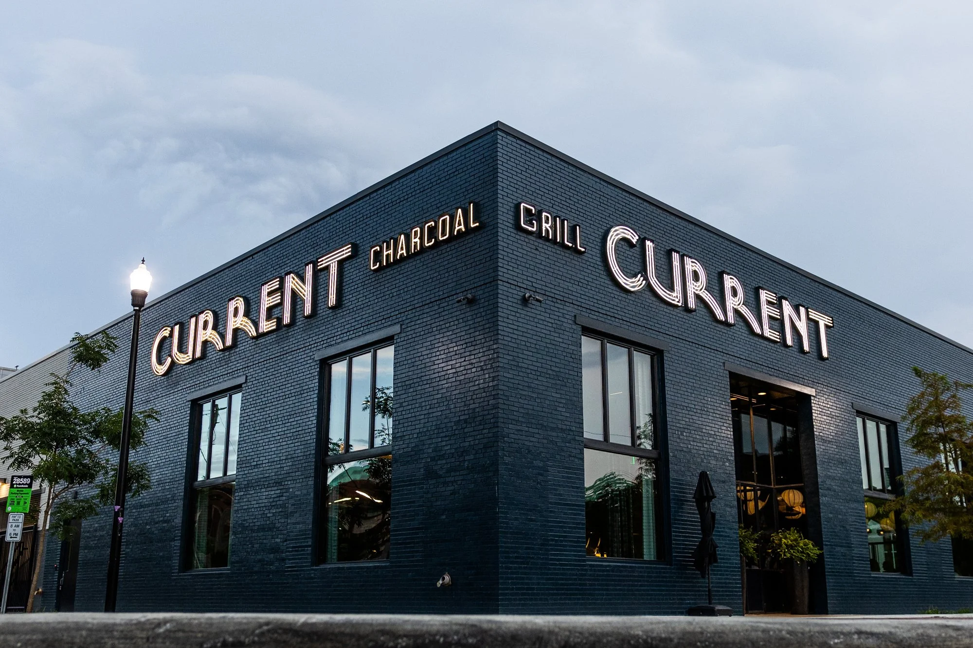 A modern dark brick building with large windows and neon signs displaying 'Current', 'Charcoal', 'Grill', and 'Cuisine' on its exterior, with street lamps and trees nearby, under a cloudy sky.
