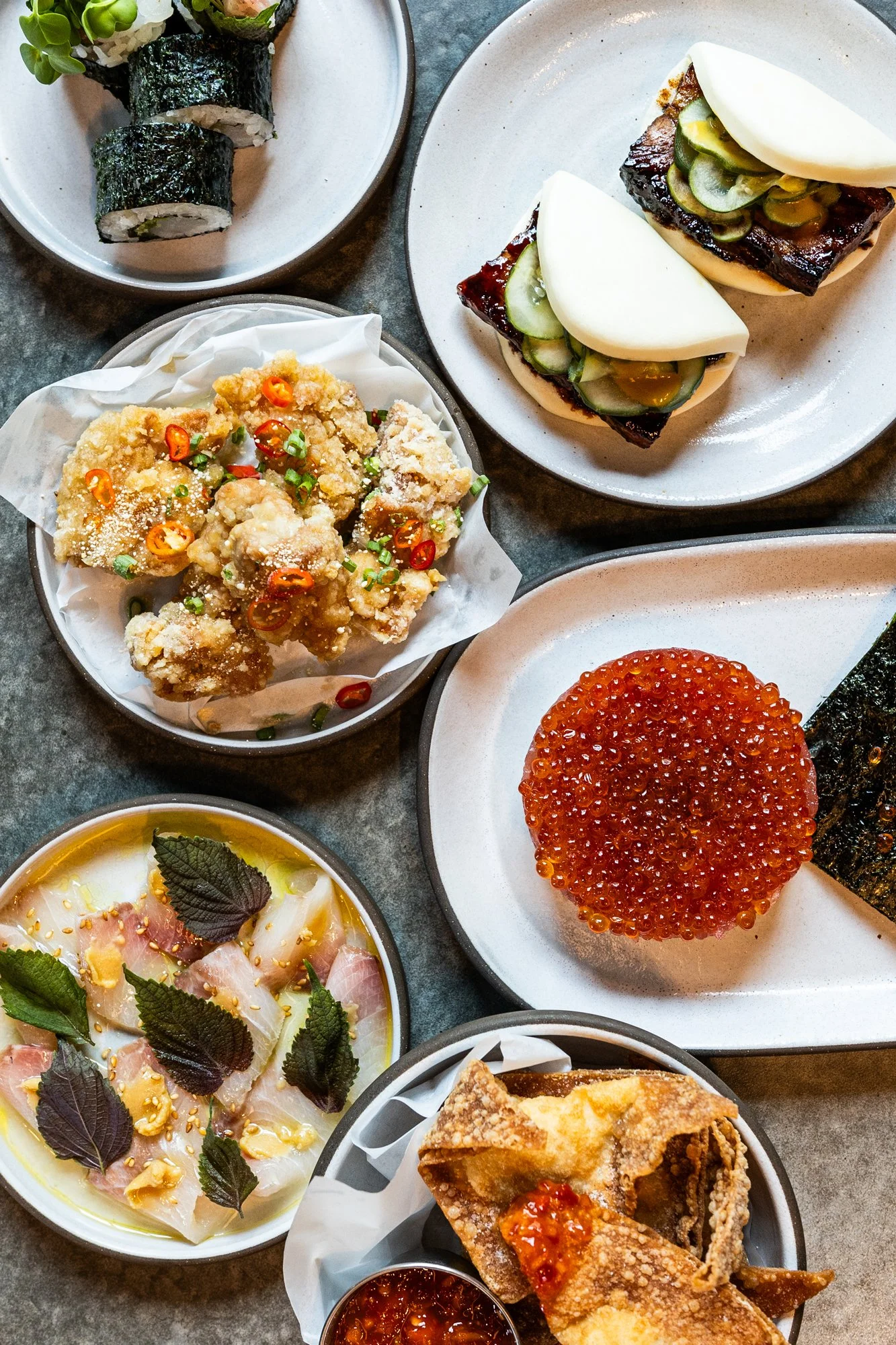 Assorted Japanese dishes on plates, including sushi rolls, fried chicken wings with chili peppers, rice balls with fish roe, and slices of sashimi garnished with herbs. Birmingham, Alabama.