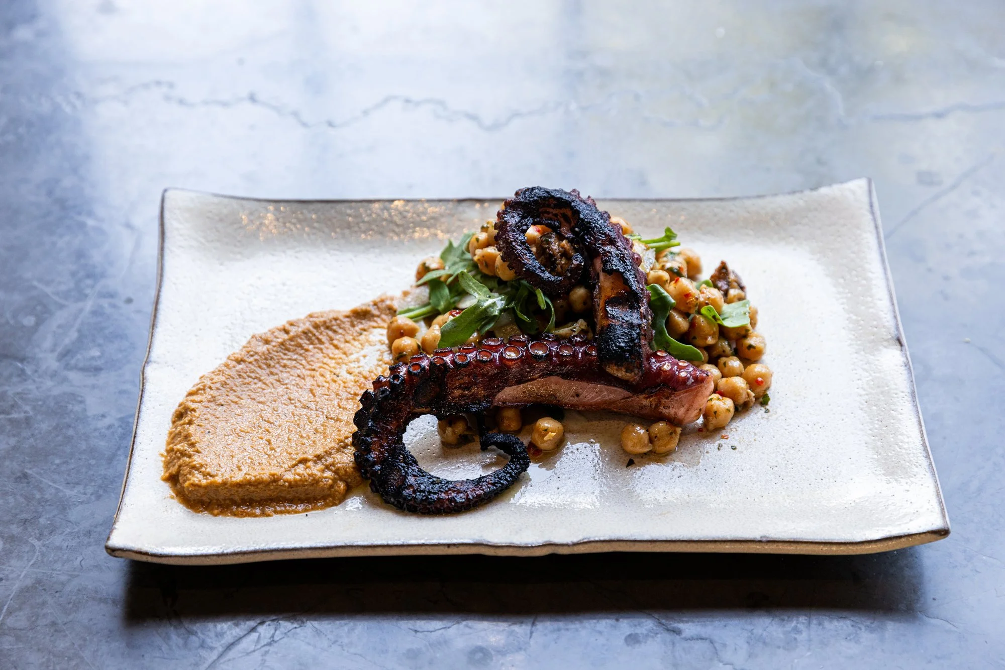 Grilled octopus tentacle served with chickpea salad and a smear of hummus on a rectangular white plate. Hot and Hot Fish Club Restaurant in Birmingham, Alabama.