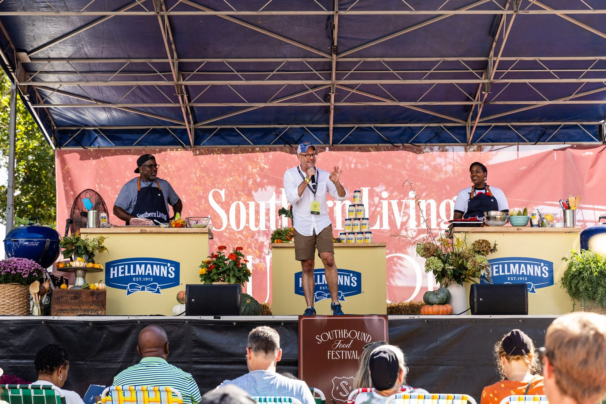 A man stands on stage speaking into a microphone at the Southbound Food Festival, with two women behind counters labeled Hellmann's on either side, under a large canopy. The stage is decorated with flowers, pumpkins, and hanging plants, and an audien