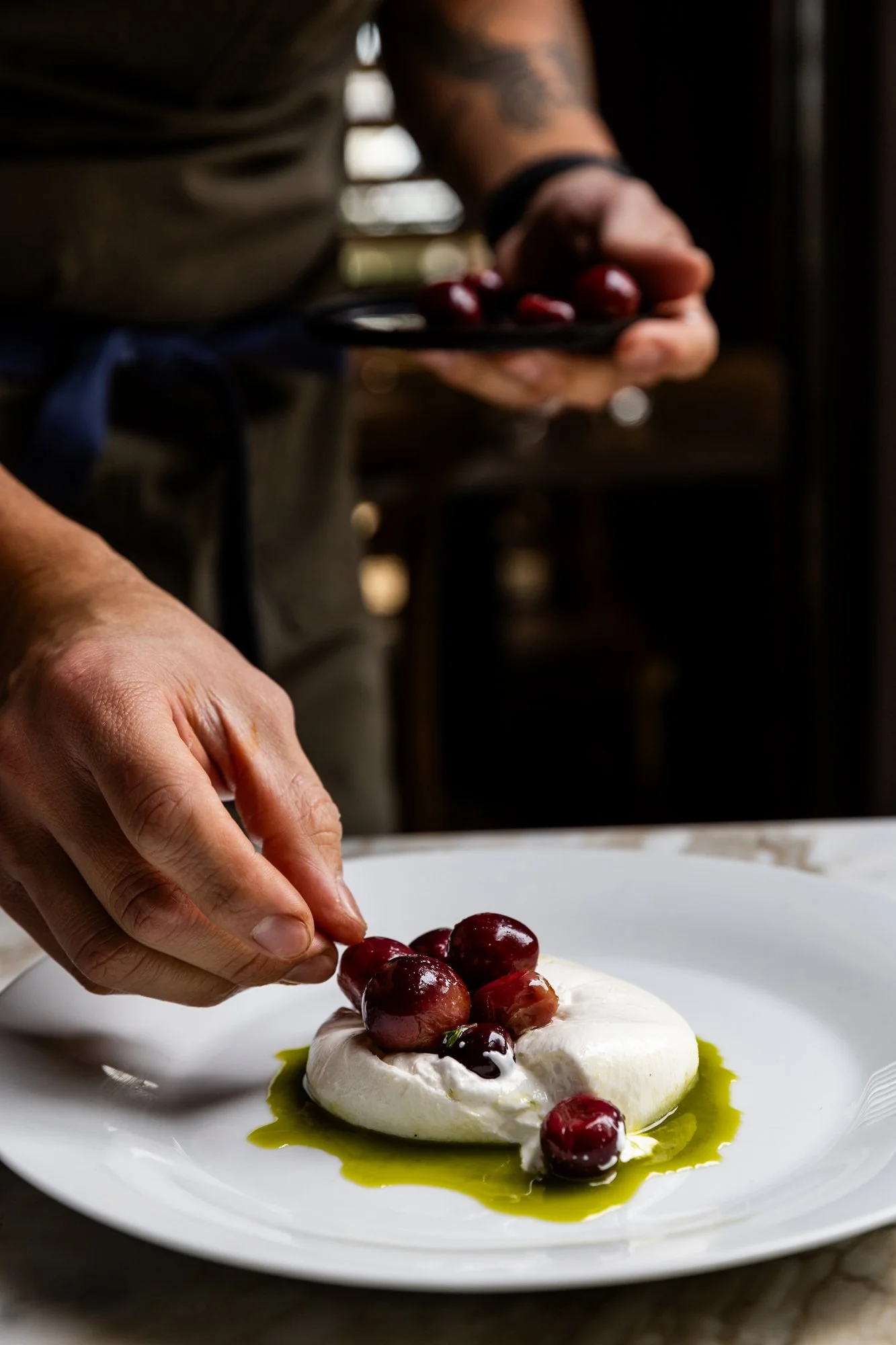 Person placing grapes on a white cheese with olive oil drizzle, seen in close-up. Ovenbird Restaurant in Birmingham, Alabama.