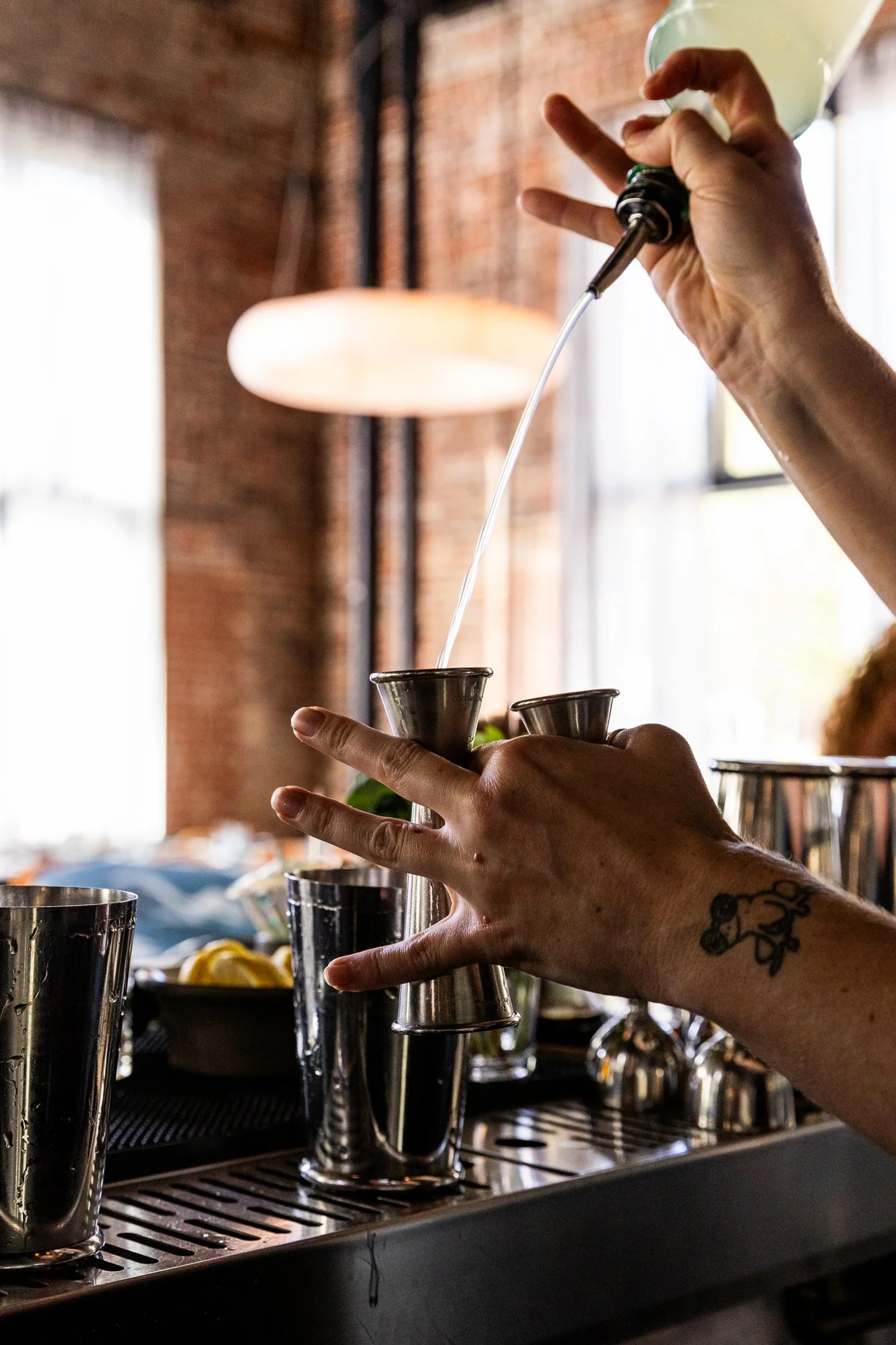 A person pouring liquid from a squeeze bottle into a metal jigger on a bar counter surrounded by cocktail shakers and bar tools, with a brick wall and window in the background.